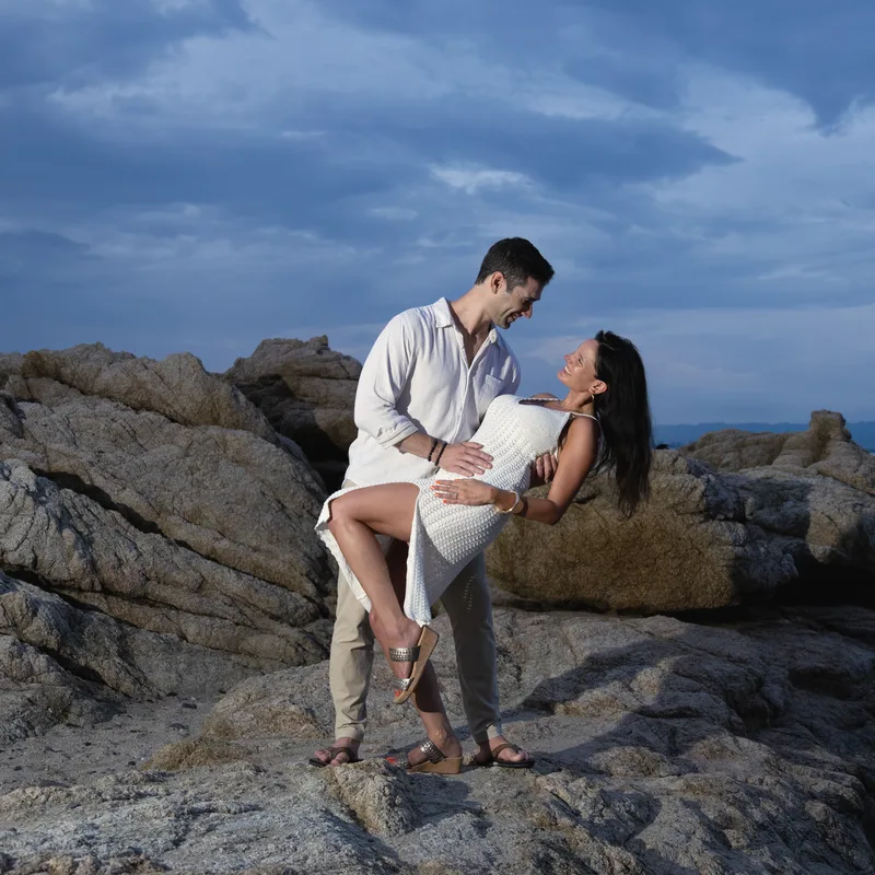 Charlie dipping Carmela on dusk-lit rocks by the sea, both laughing, after the proposal