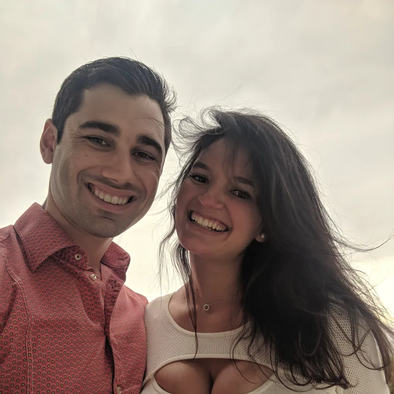 Carmela and Charlie laughing in a windy close-up selfie against a pale sky