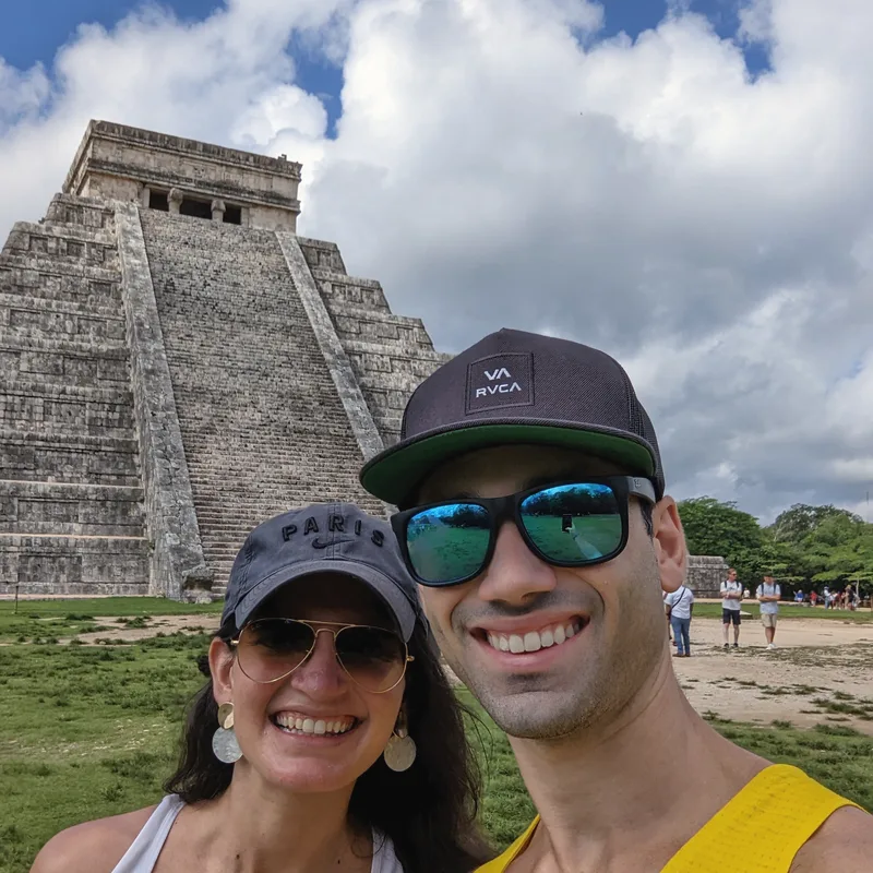 Carmela and Charlie smiling in a selfie in front of the stepped stone Mayan pyramid at Chichen Itza