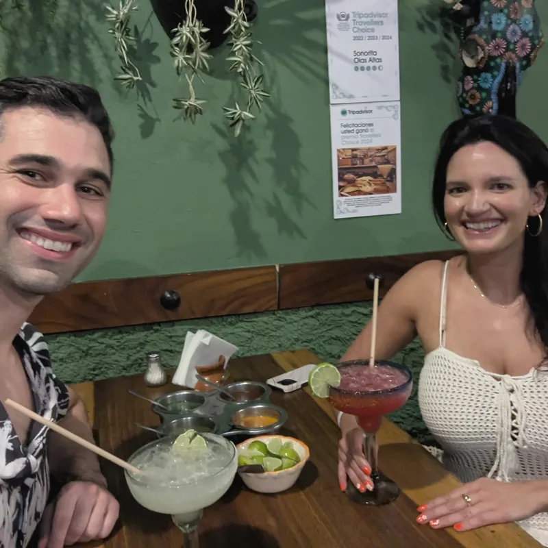 Charlie and Carmela at a restaurant table with margaritas, a green wall behind them