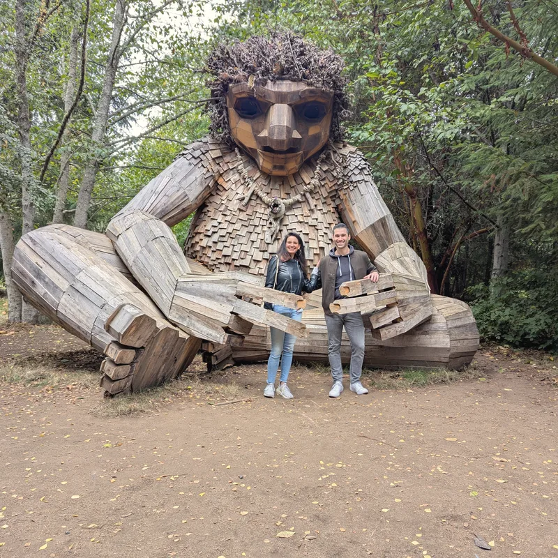 Carmela and Charlie standing in front of a giant wooden troll sculpture in a forest clearing