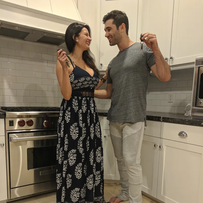 Carmela and Charlie in a kitchen, each holding up a set of new keys and smiling at each other