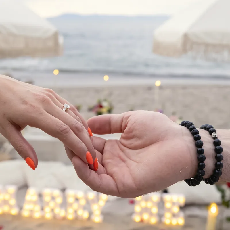 A hand with an engagement ring resting in another hand, with softly lit marquee letters spelling marry me on the sand