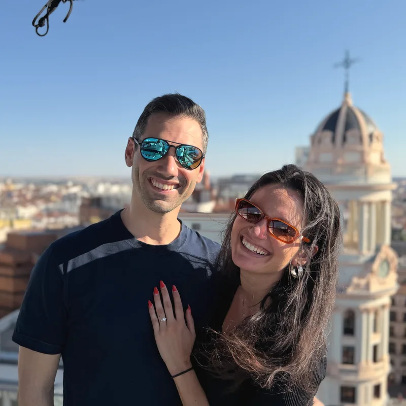 Charlie and Carmela in sunglasses smiling on a rooftop, with a domed Madrid skyline behind them