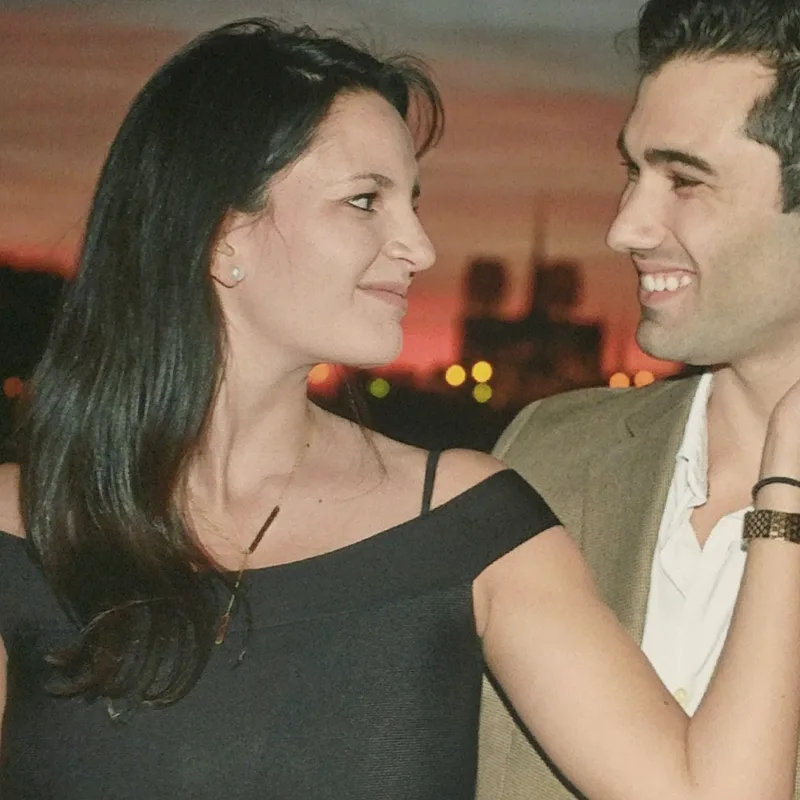 Carmela and Charlie sharing a warm look at sunset in an early photograph, city lights glowing behind