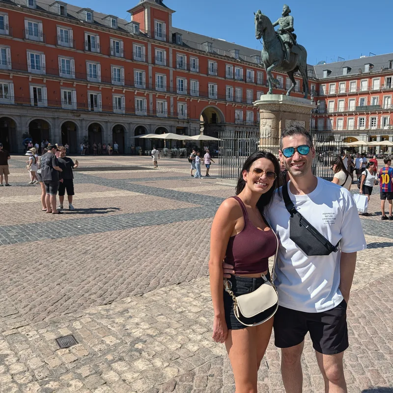 Carmela and Charlie standing together in the sun in a cobbled plaza ringed by red-brick arcades
