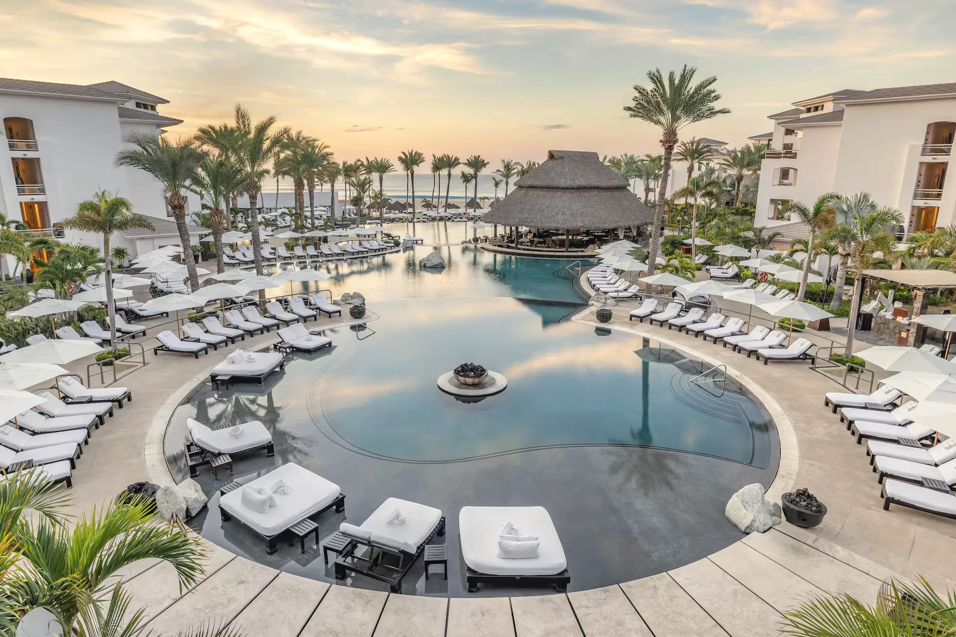Curved resort pool at sunrise, ringed by white loungers and palms, with a thatched-roof bar at the far side and white buildings around it.