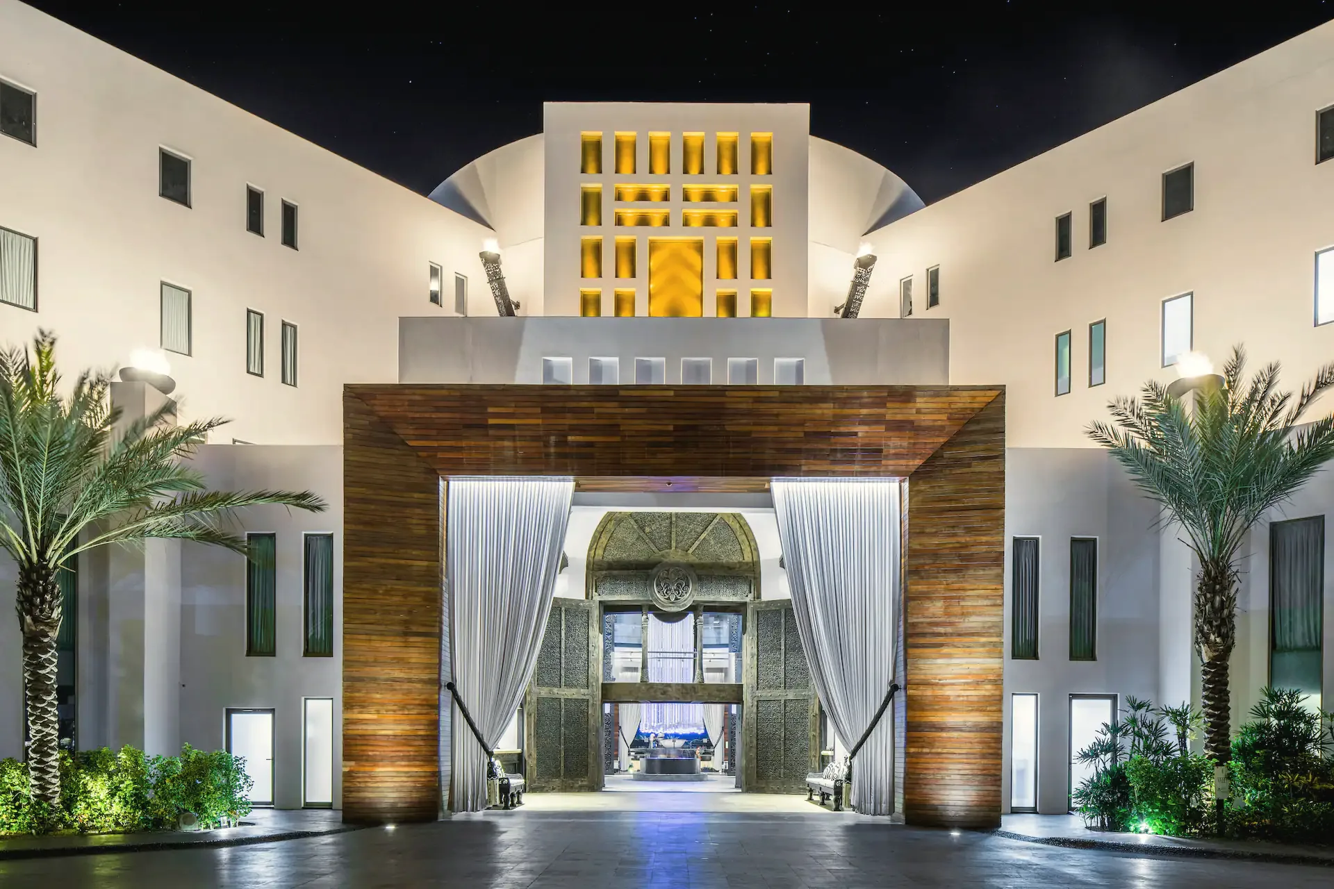 Hotel entrance at night: a timber-framed gateway with draped white curtains, a lit gold facade above, palms and landscape lighting.