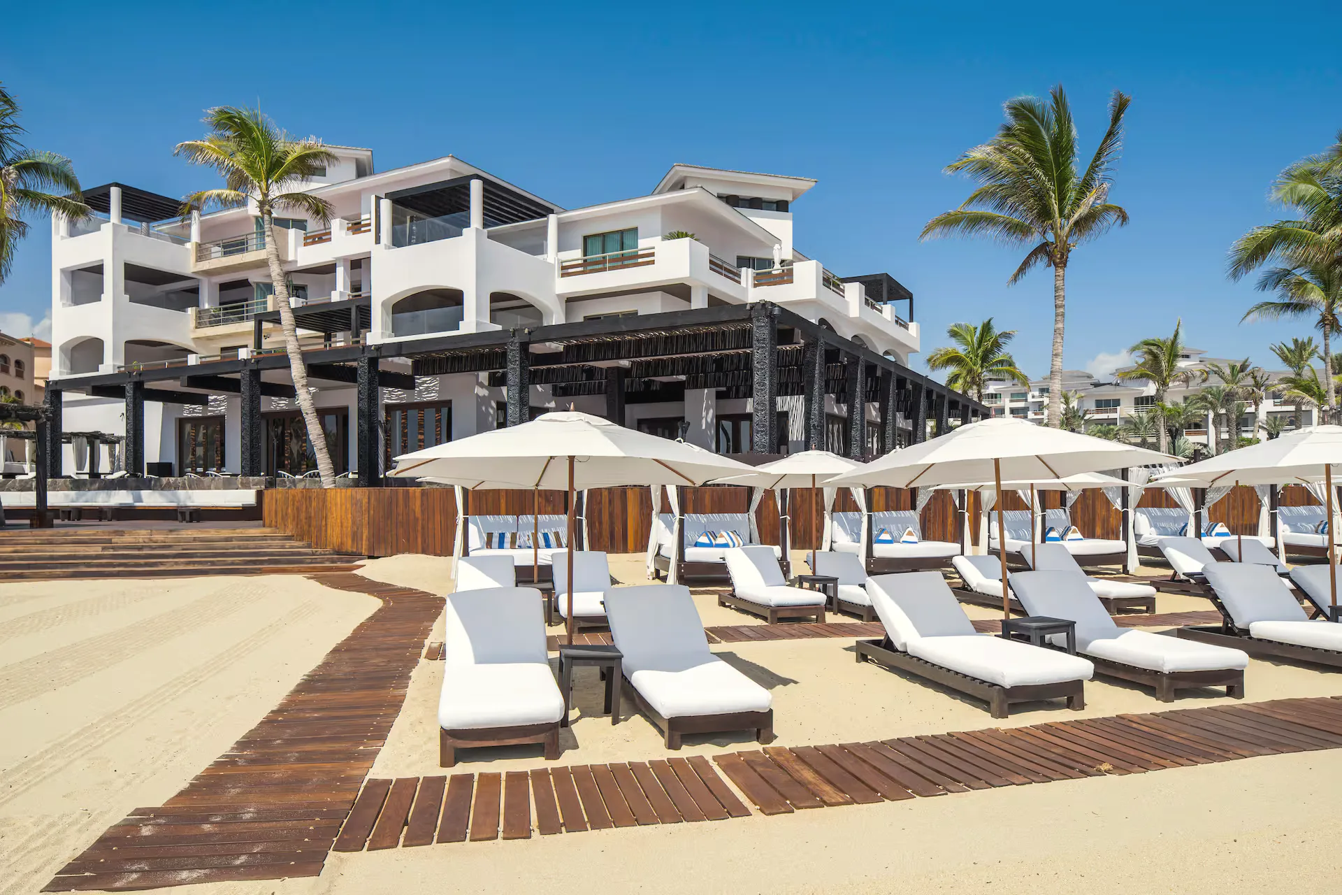 White loungers and umbrellas on the sand beside a dark timber boardwalk, with a white multi-storey building behind.