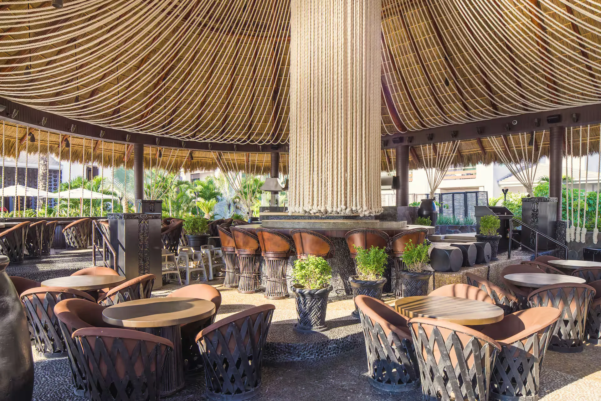 Open-air lounge under a high woven-rope ceiling, with round tables and leather-strapped chairs around a central column.