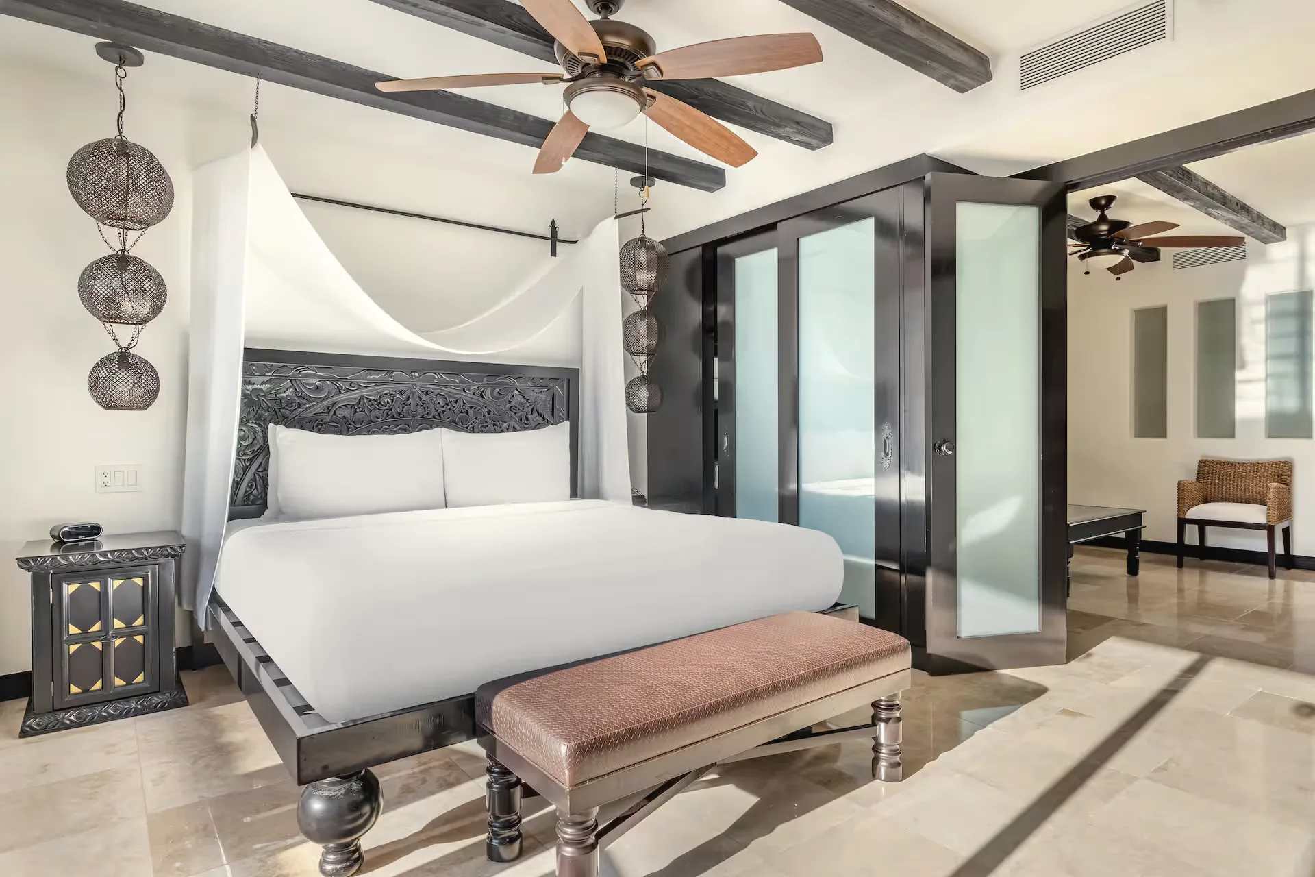 Bedroom with a dark carved headboard, white bed linens with a draped canopy, a leather bench at the foot of the bed and a ceiling fan.
