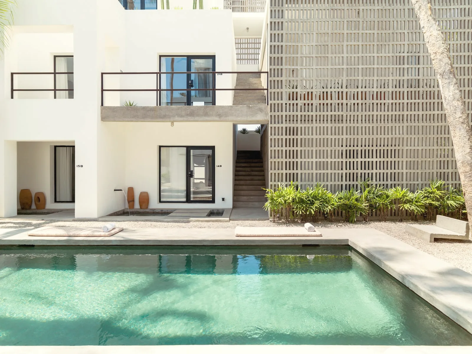 Small rectangular pool with turquoise water in front of a white two-storey building and a tall concrete screen wall, with a palm trunk at the right.