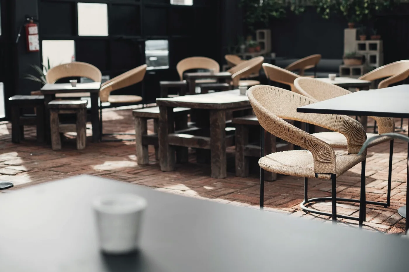Outdoor seating on a brick floor with dark wooden tables and woven chairs, with a glass in sharp focus in the foreground.
