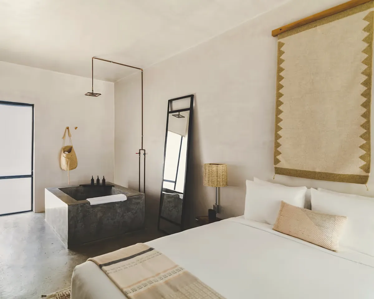Bedroom corner with a white bed, a woven wall hanging, a dark tiled bathtub and a copper shower pipe.