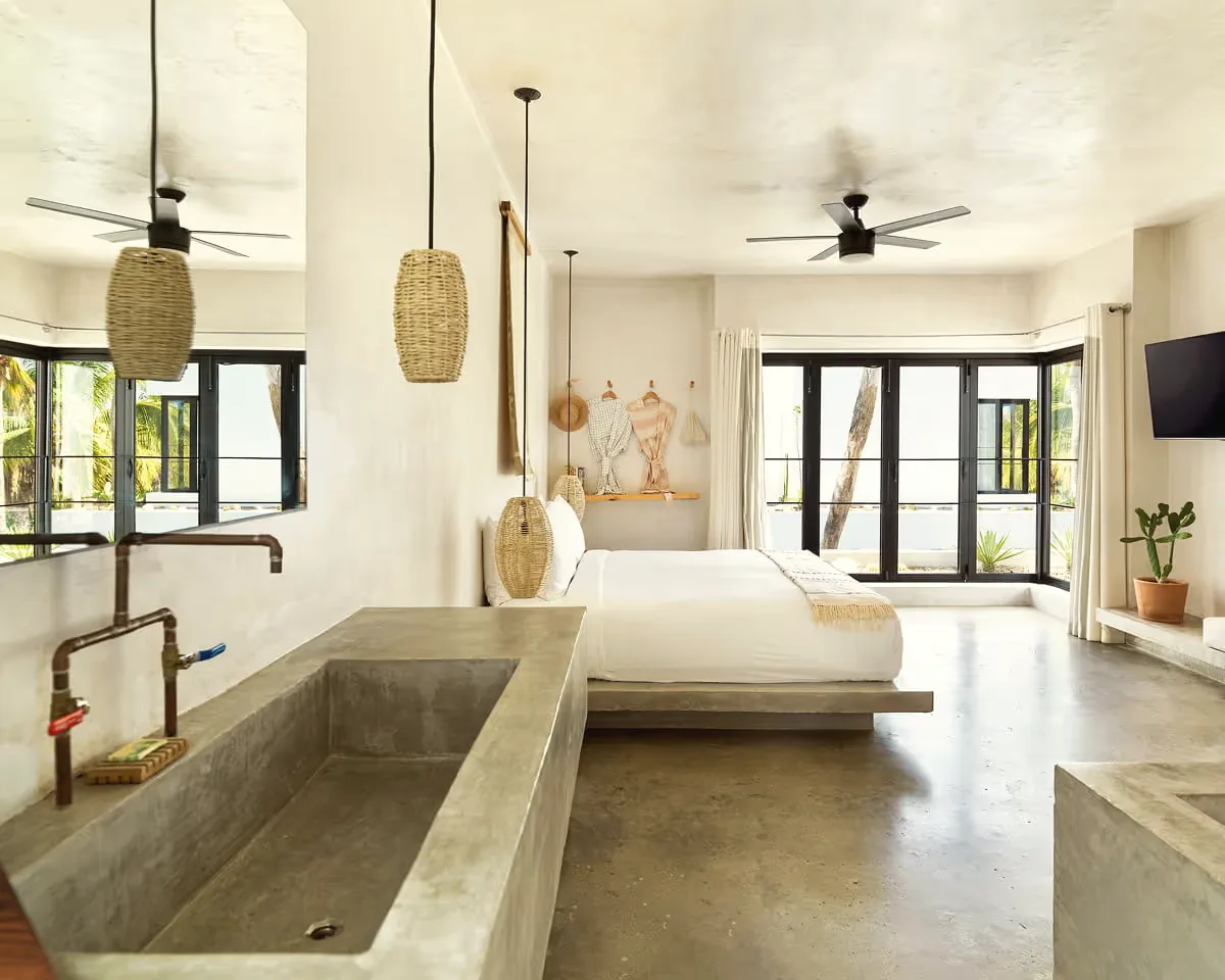 Room with a long concrete trough sink with copper taps in the foreground, a low white bed behind it and windows onto greenery.