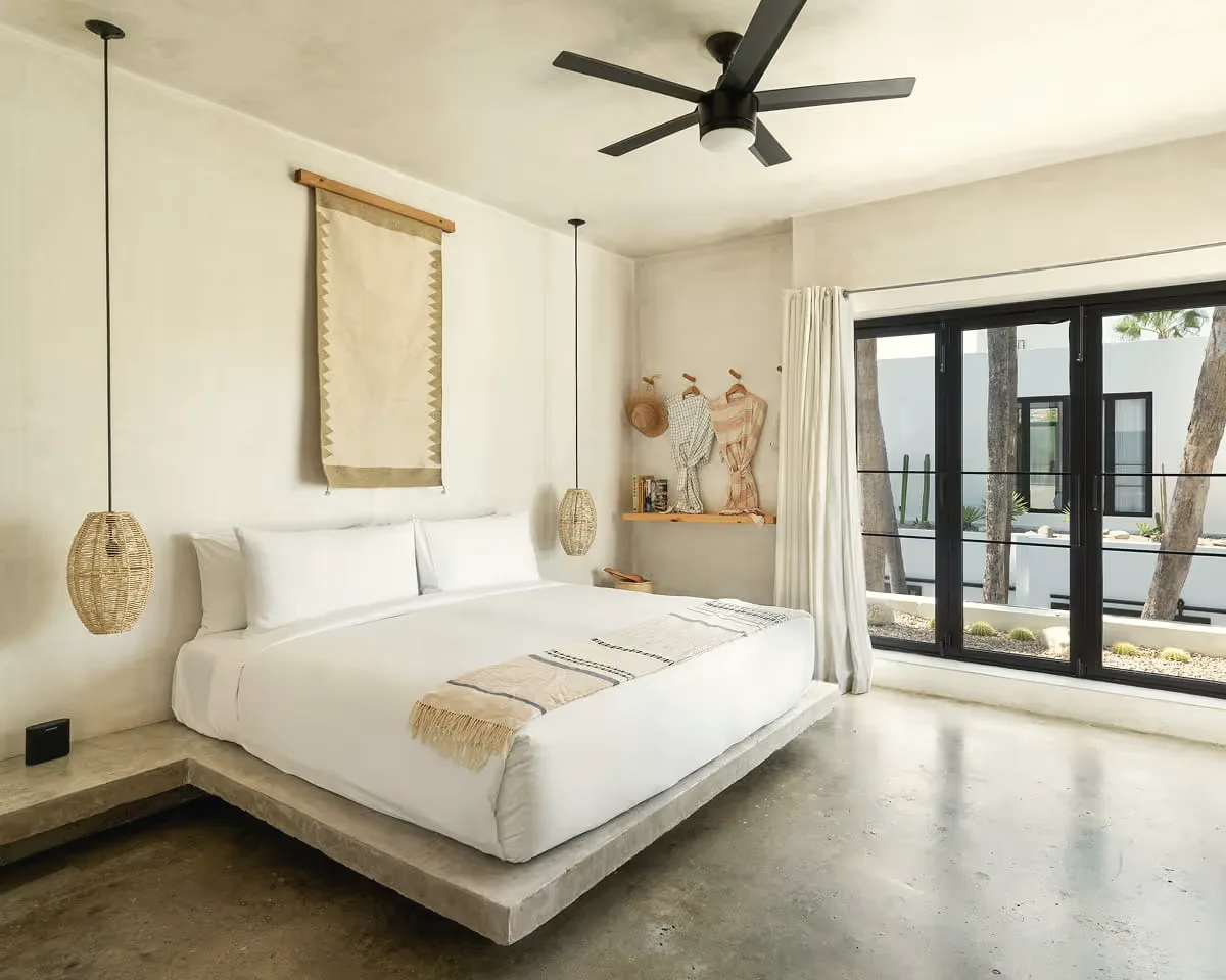 Bedroom with a low white bed, a woven wall hanging, rattan pendant lamps, a ceiling fan and black-framed glass doors.
