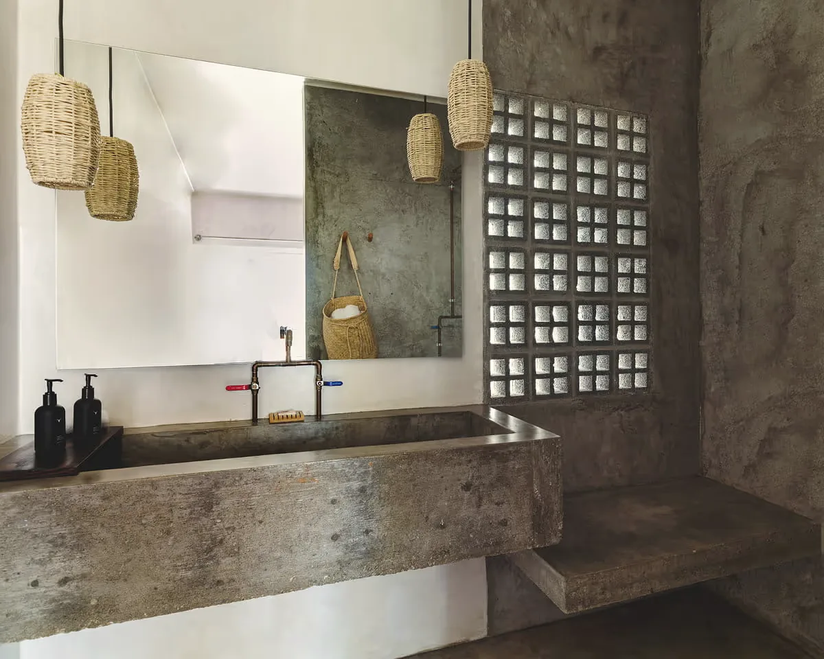 Washbasin carved from concrete with copper taps, a mirror, rattan pendant lamps and a glass-block window beside a textured grey wall.