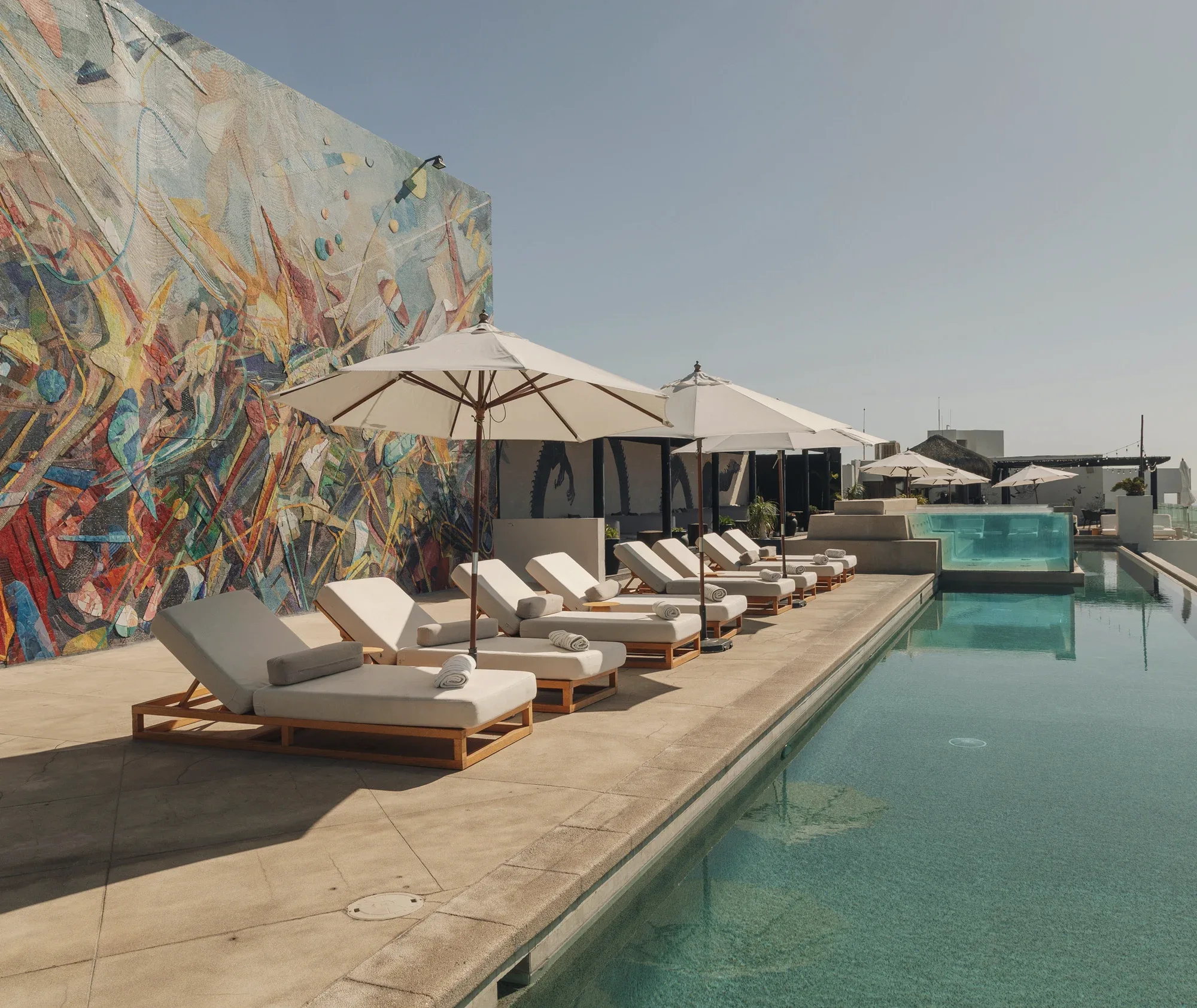 Rooftop terrace with a row of cushioned wooden loungers and cream umbrellas between a large colourful abstract mural and a long pool with a glass-sided end.