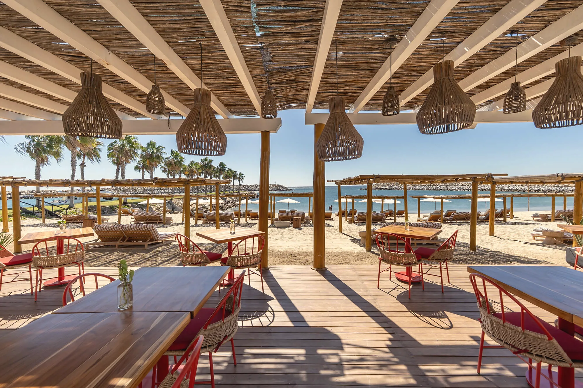 Covered wooden deck with tables and red chairs under a reed ceiling with woven pendant lamps, looking out over beach loungers toward the sea.
