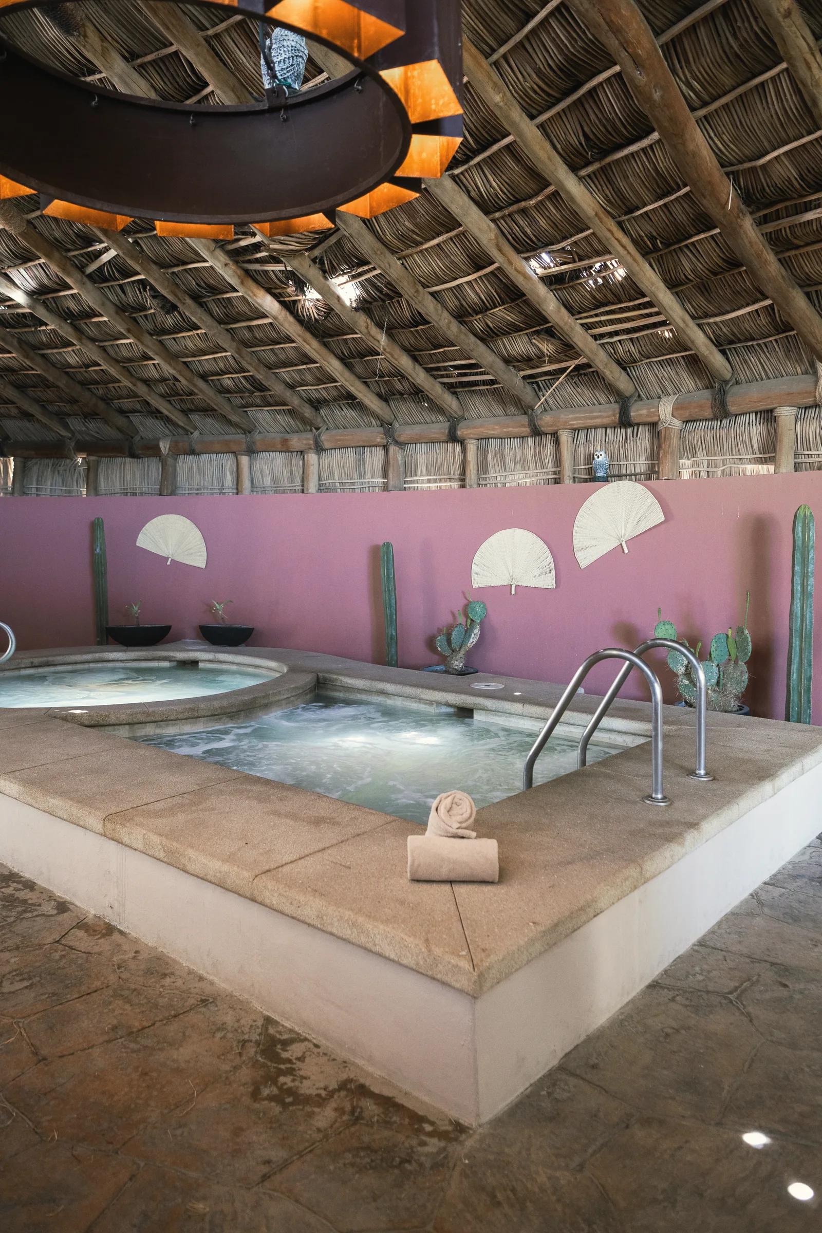 Stone-edged pool with bubbling water under a thatched palm-leaf roof, with a rolled towel on the rim, a pink wall and potted cacti.