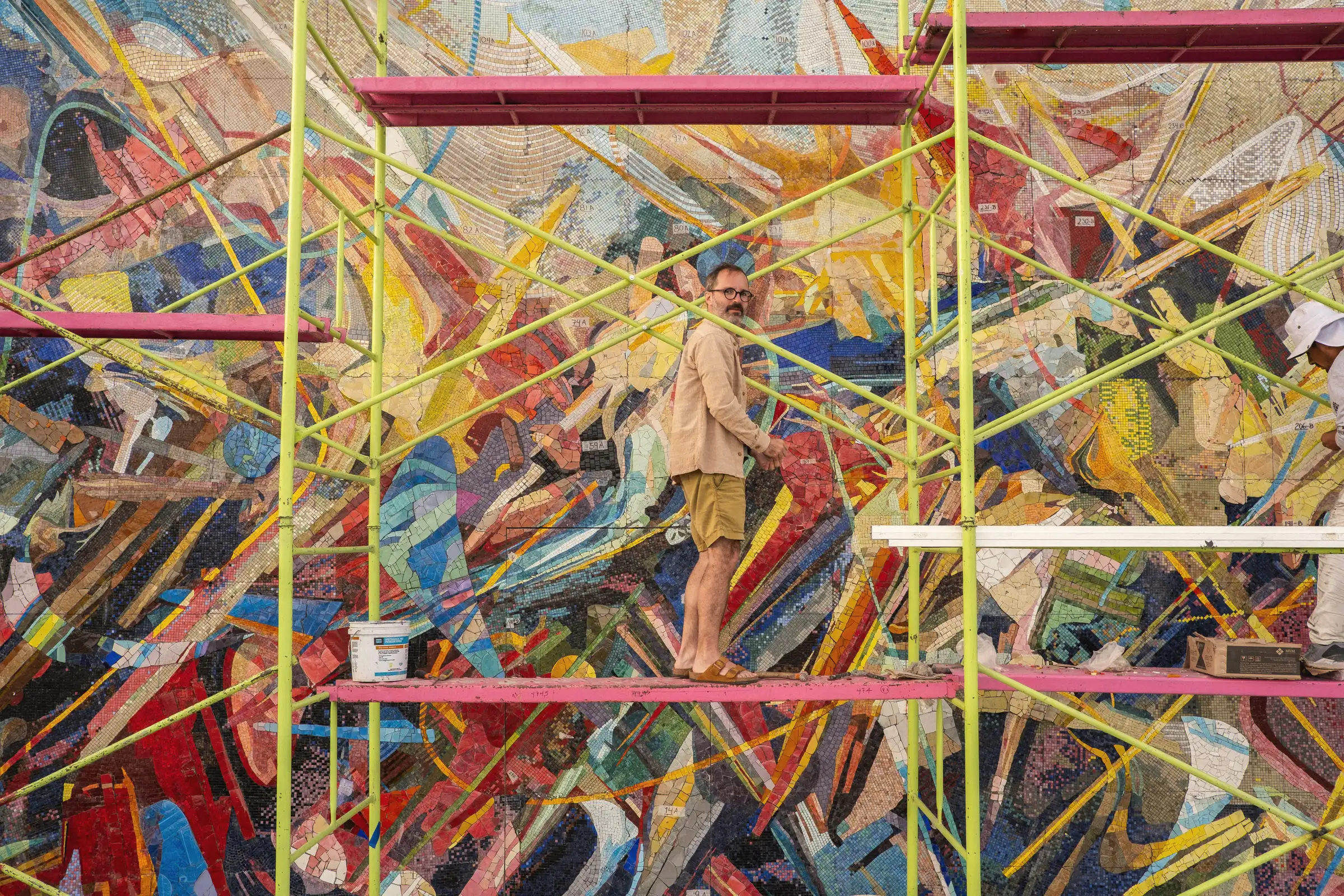 A man standing on bright pink and yellow-green scaffolding in front of a huge mosaic mural of colourful angular fragments.