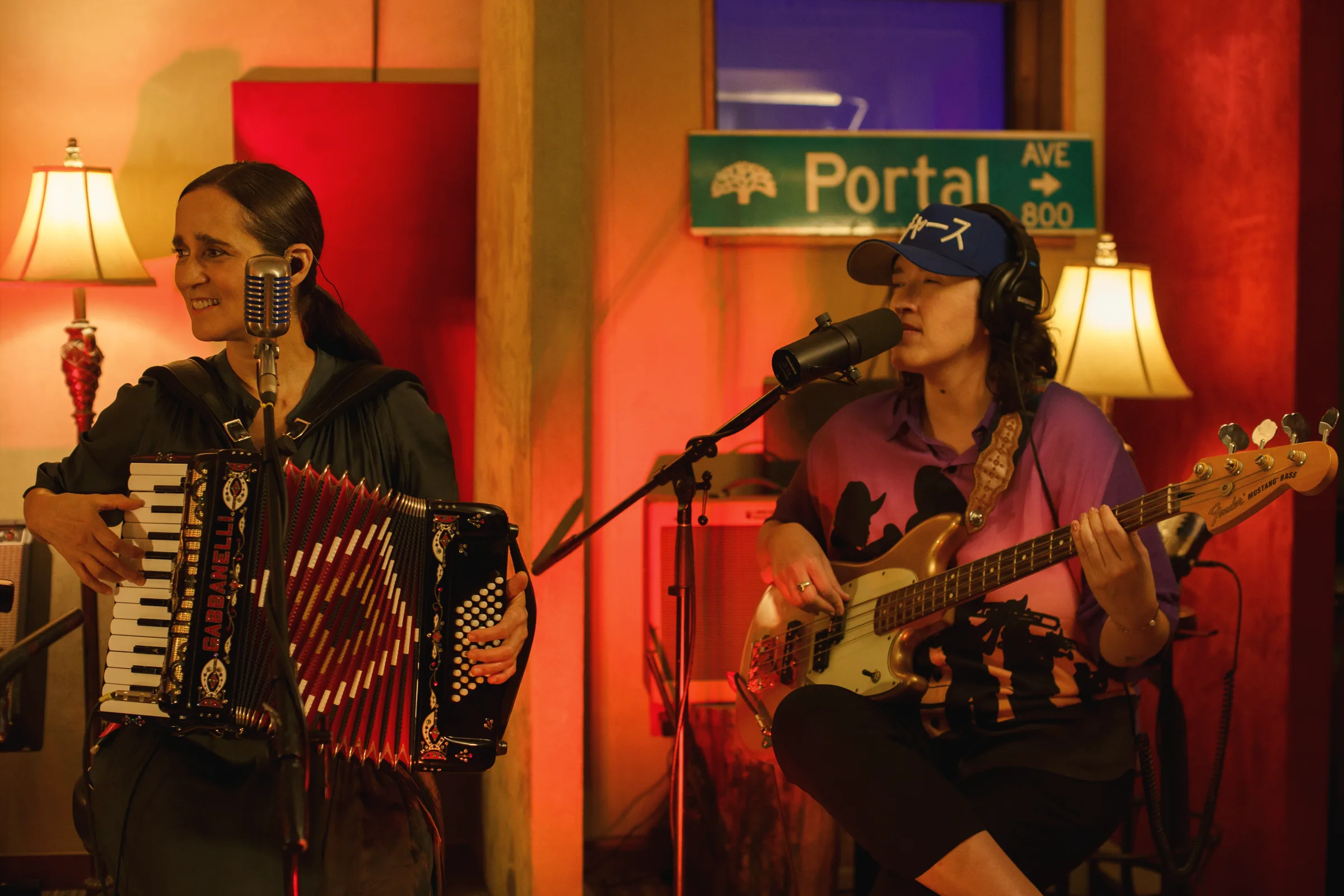 Two musicians performing in a warmly lit red room, one singing and playing an accordion, the other singing and playing a bass guitar, under table lamps and a street-sign style Portal sign.