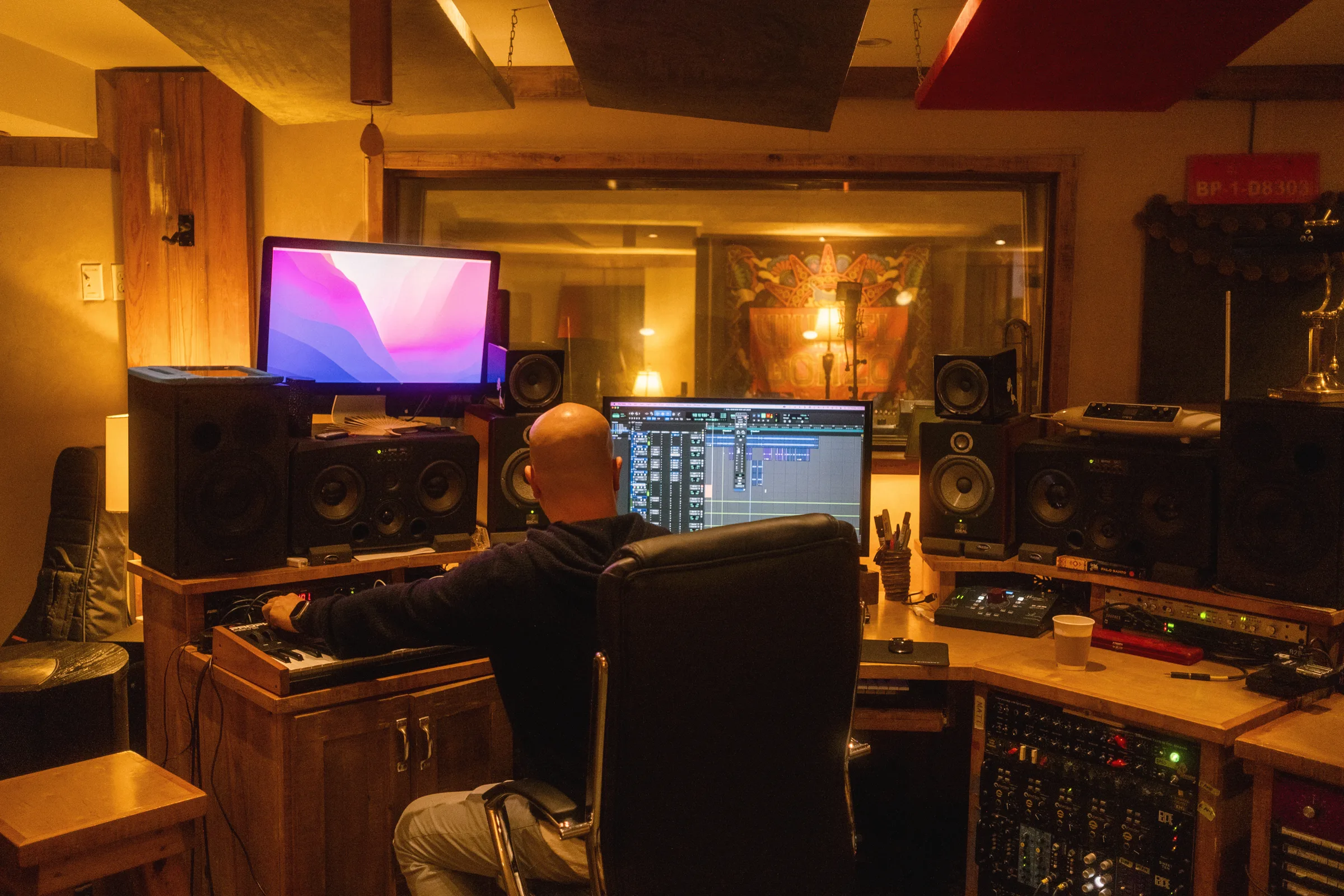 A man seated at a recording console with two computer screens and studio monitors in a warmly lit wood-panelled studio.