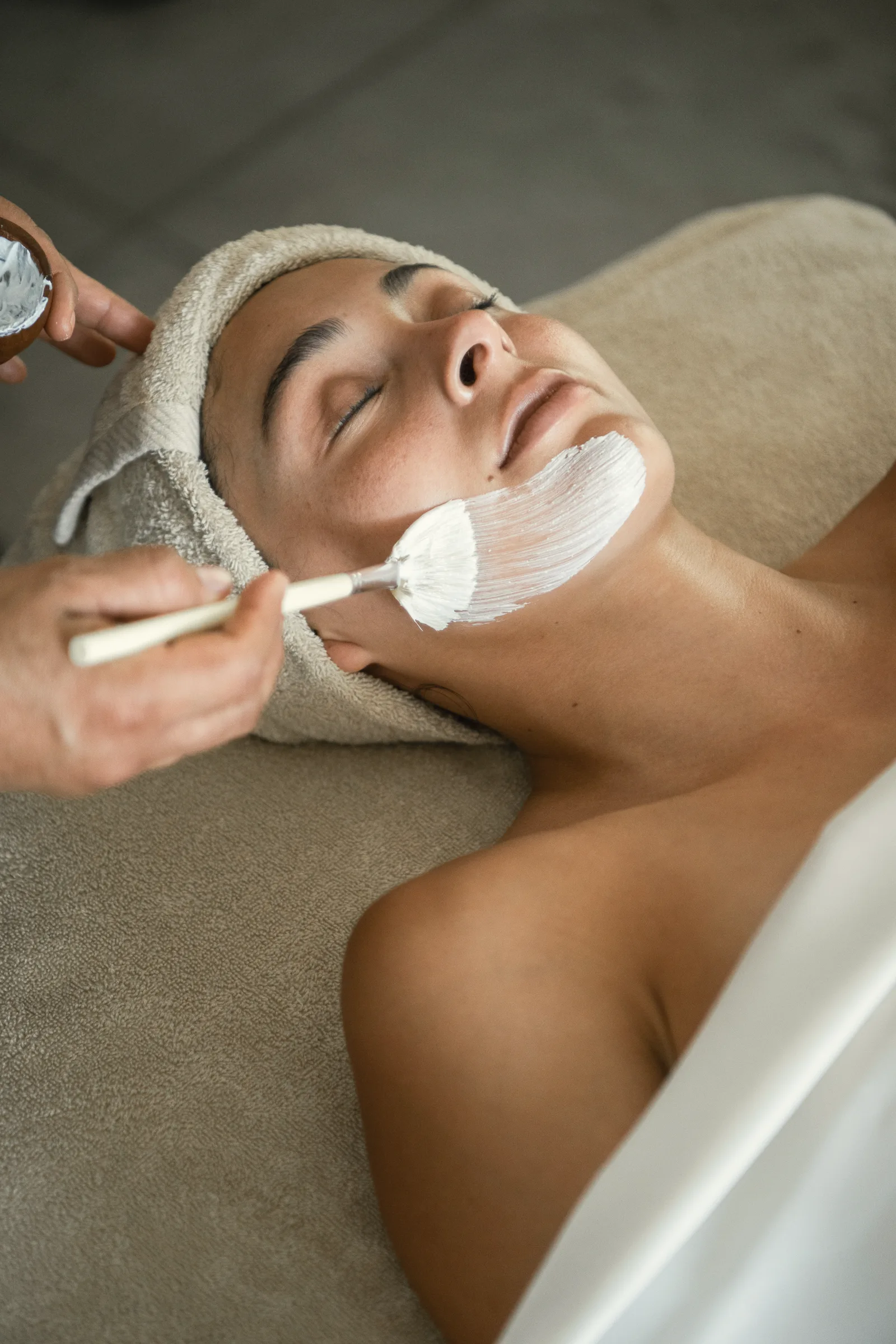 Close view of a guest lying back with eyes closed, wearing a head towel while a white face mask is brushed on.