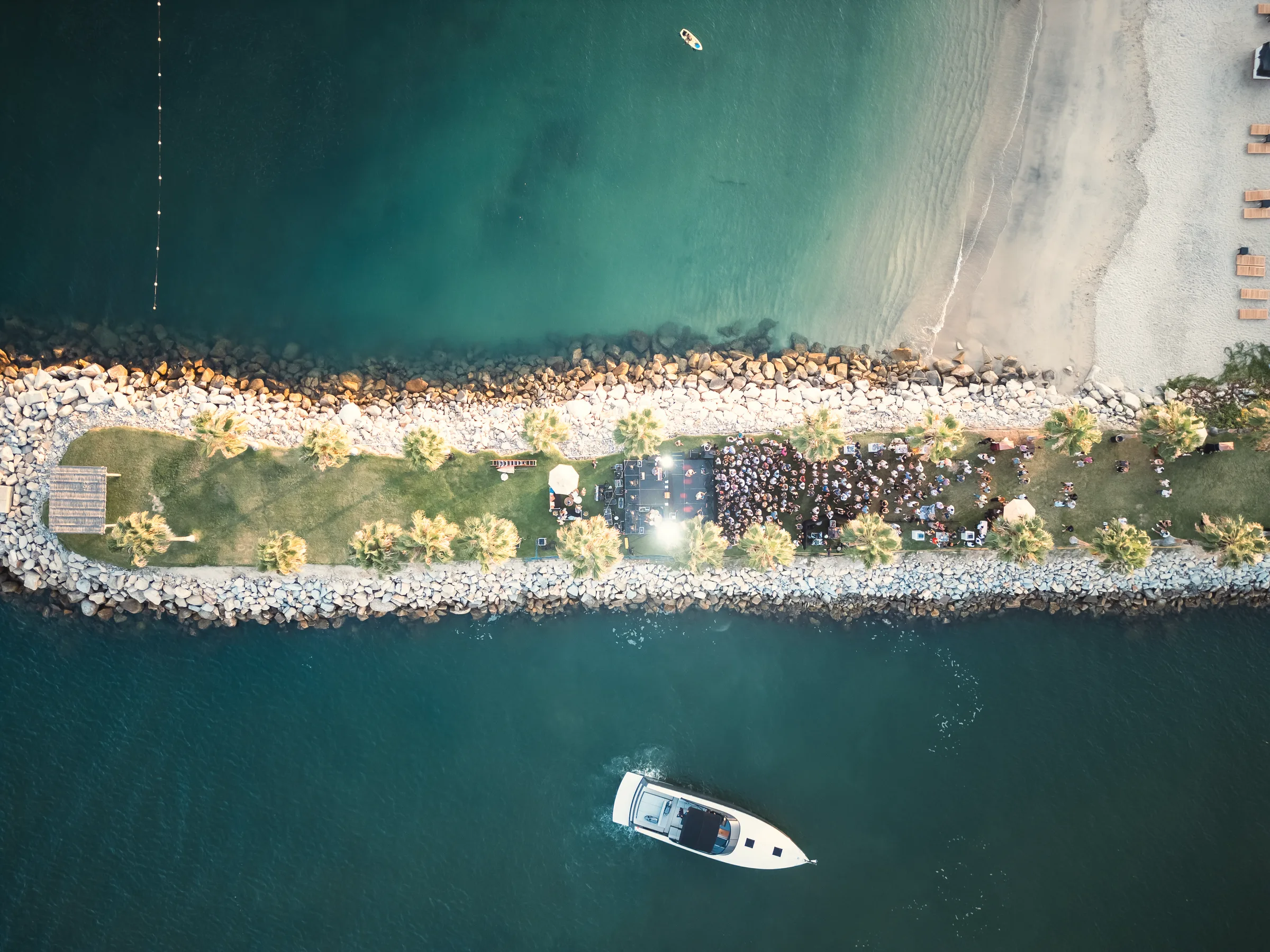 Aerial view of a narrow rock-lined jetty with palms and a crowd gathered at one end, between turquoise water and the beach, with a small boat below.