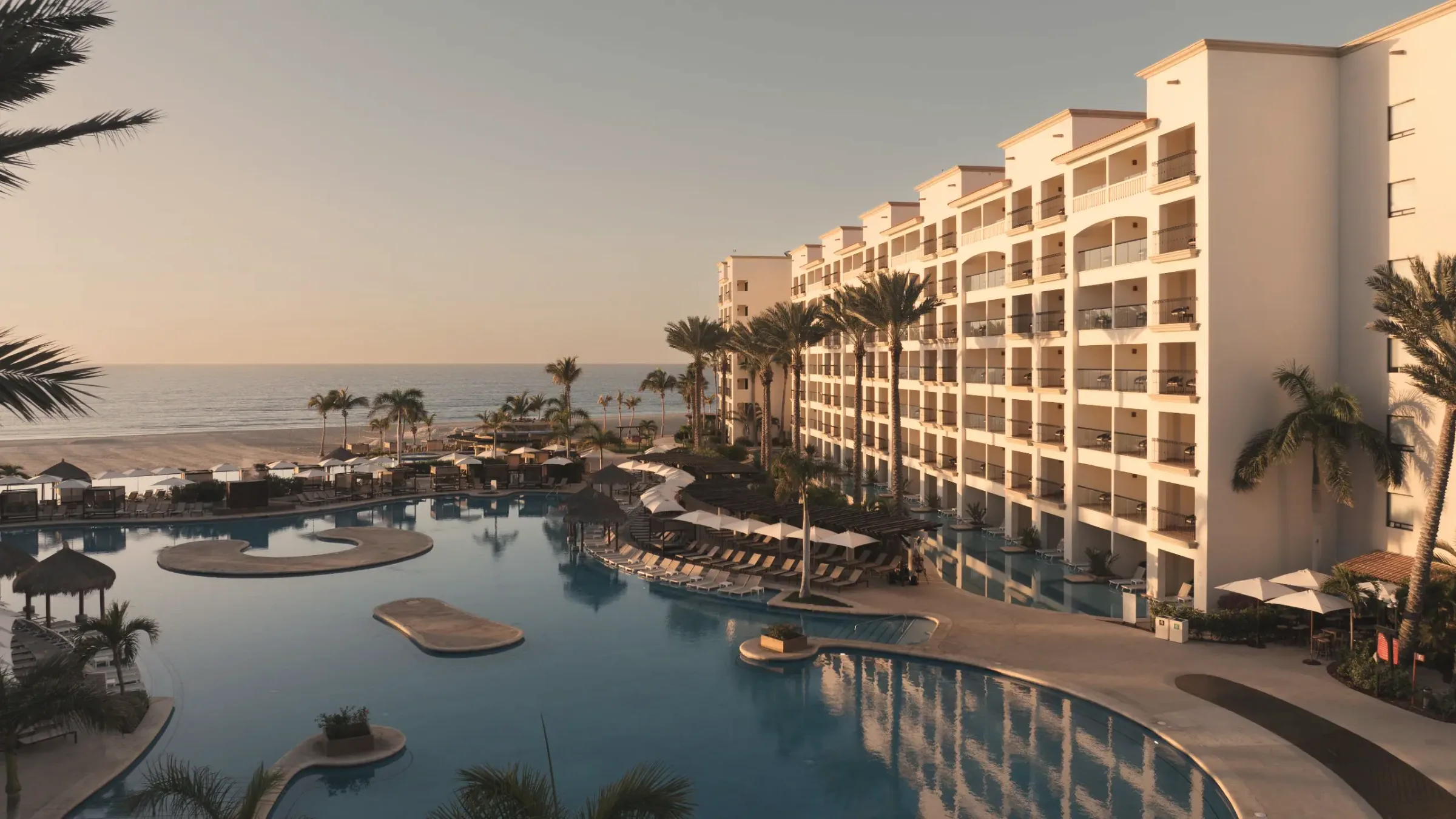 Pool terraces at golden hour, with curved pools and sandy islands in front of a long cream-coloured terraced hotel building, and the beach and sea beyond.