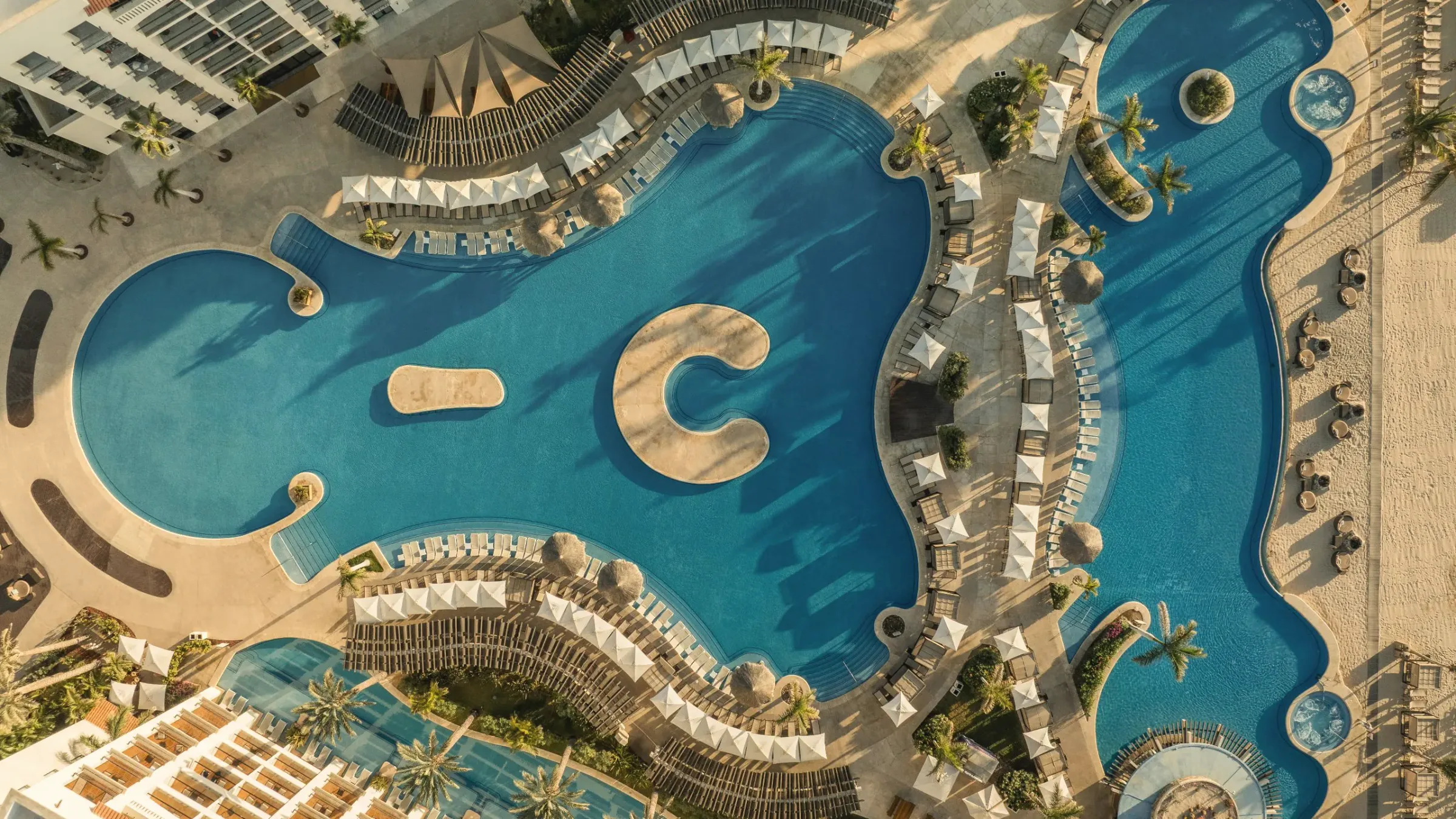 Aerial view straight down on a large curving blue pool with a C-shaped sandy island, white umbrellas and loungers around its edge, and the beach along the right side.