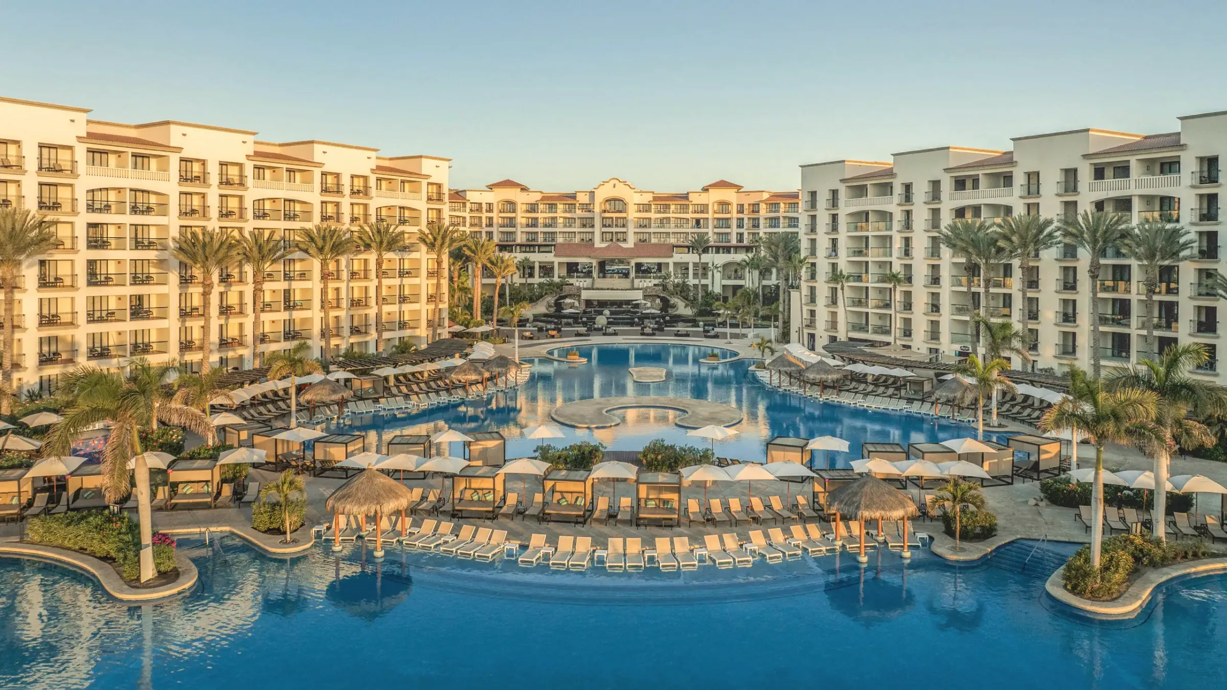 Courtyard pool framed by two cream-coloured hotel buildings, with palms, thatched shade huts, cabanas and rows of loungers around the water.