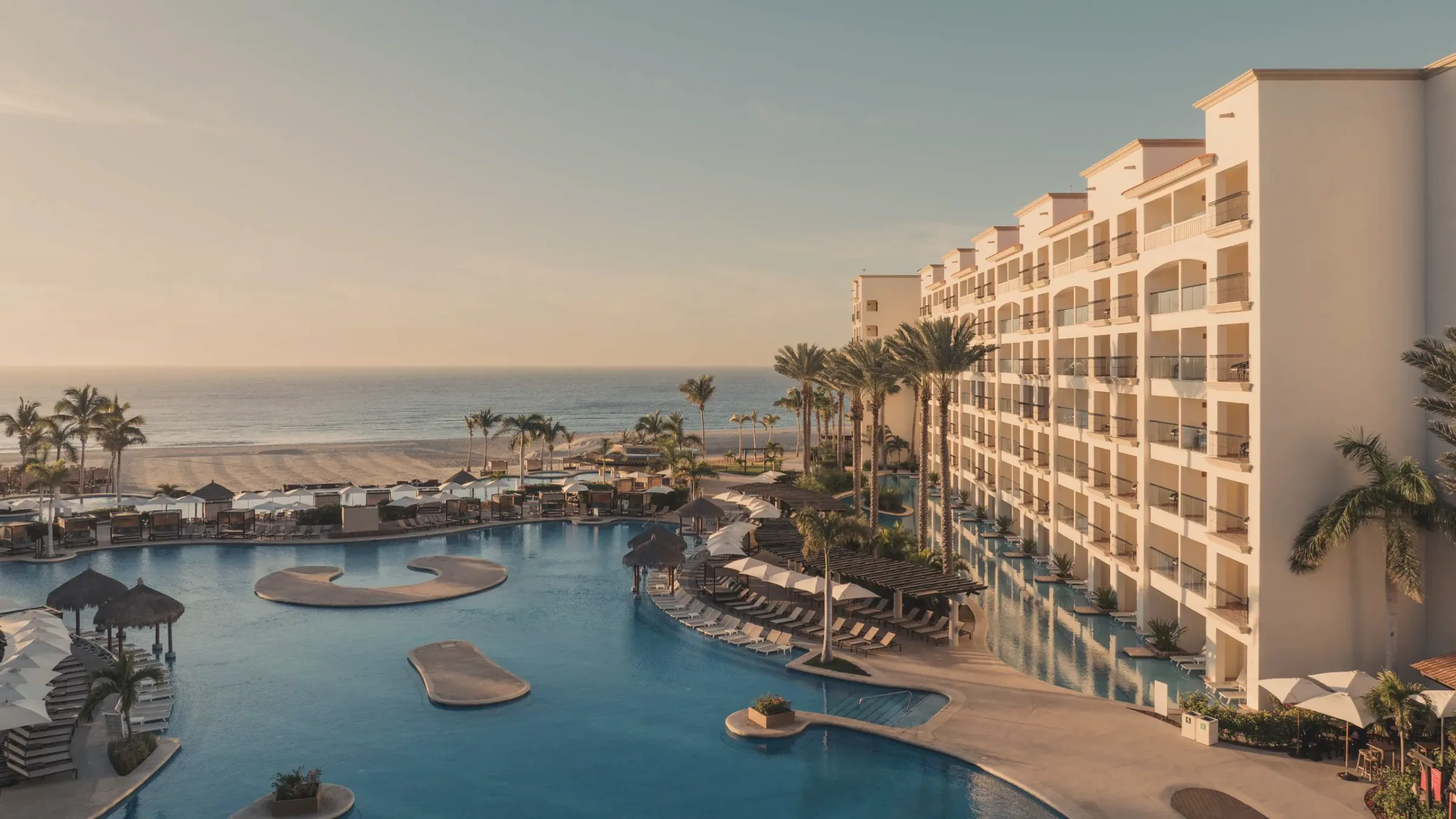 Pool deck at sunset with a curving pool, sandy islands and thatched huts, beside a tall terraced hotel building, with the beach and sea in the distance.