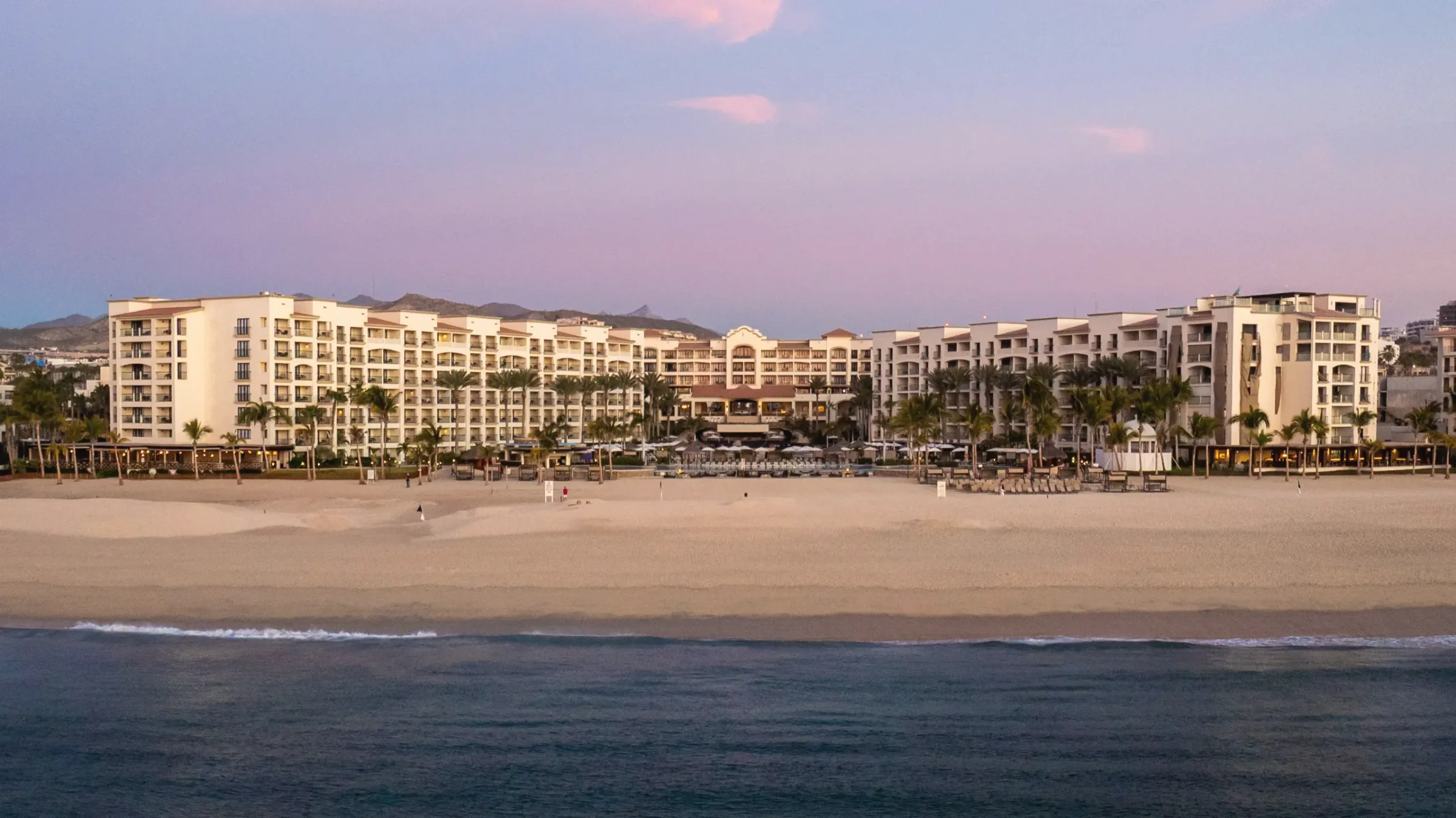 Hotel seen from the edge of the water at dusk: a long cream-coloured building with palms along an empty sandy beach, under a pink and purple sky with mountains behind.
