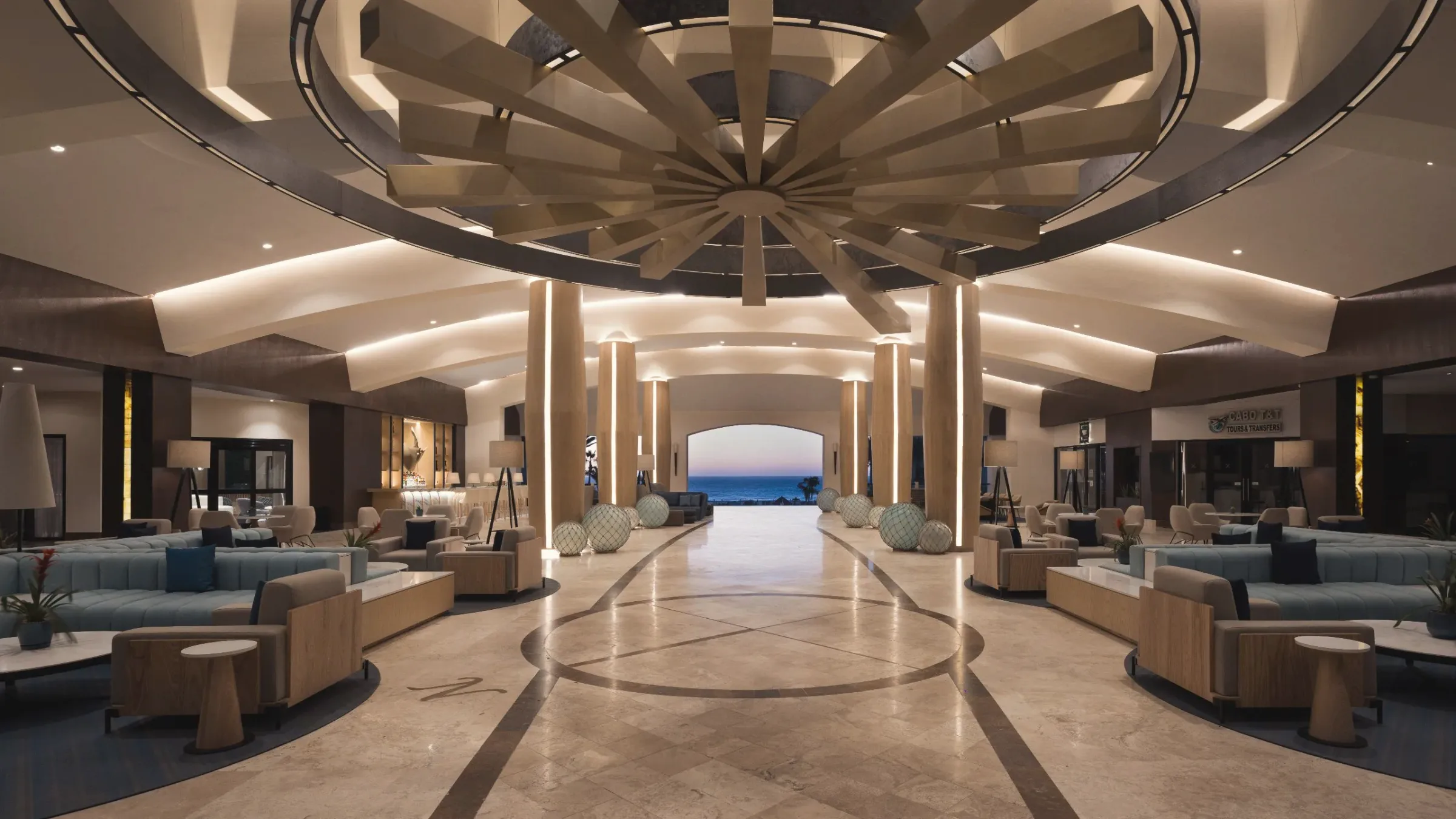 Wide lobby with a circular timber ceiling feature, pale blue sofas and wooden armchairs on both sides, and a marble floor leading to an arched opening onto the sea.