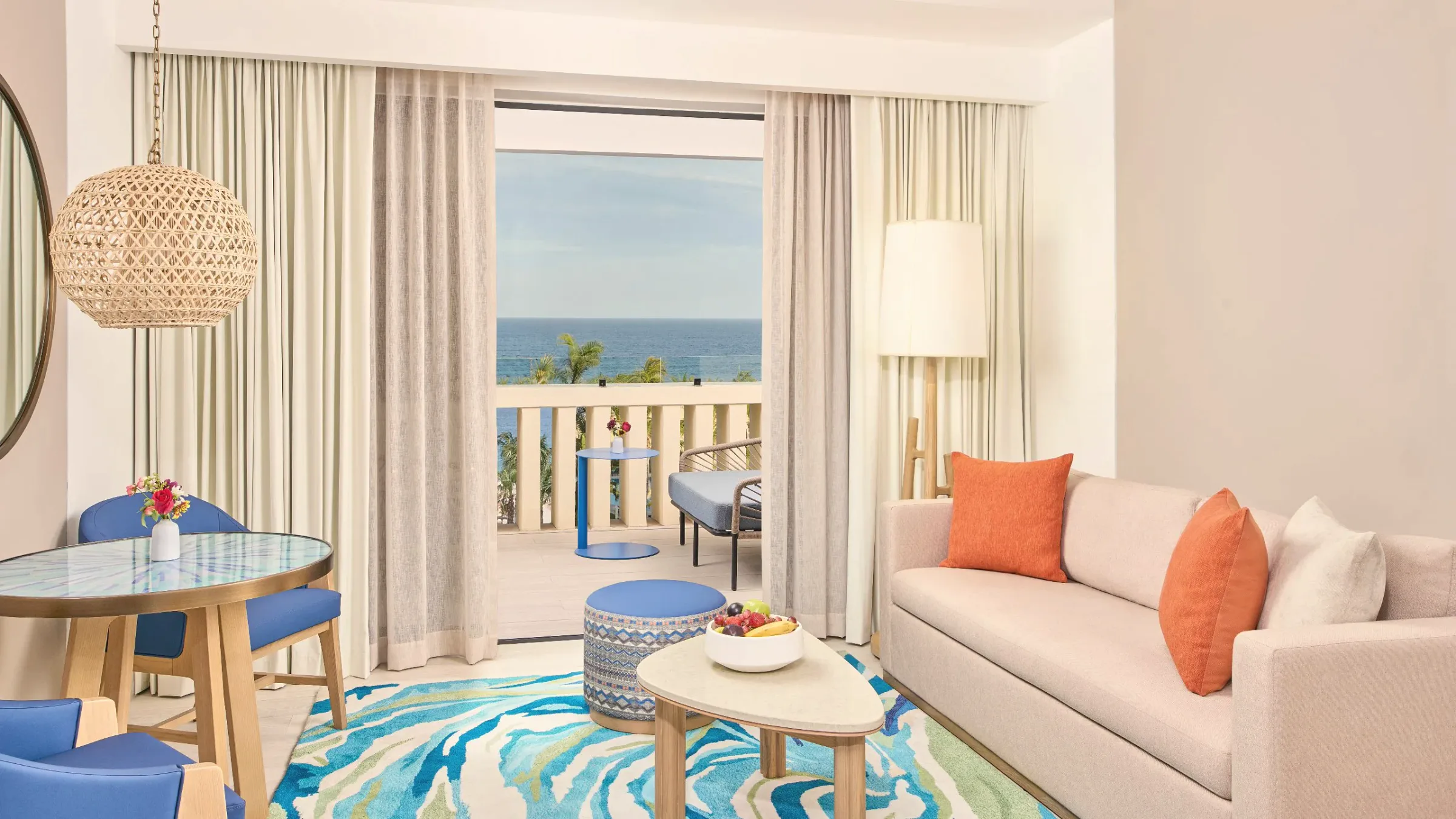 Guest room sitting area with a cream sofa with orange cushions, a round table with blue chairs and a glass door onto a balcony with a view of the sea.