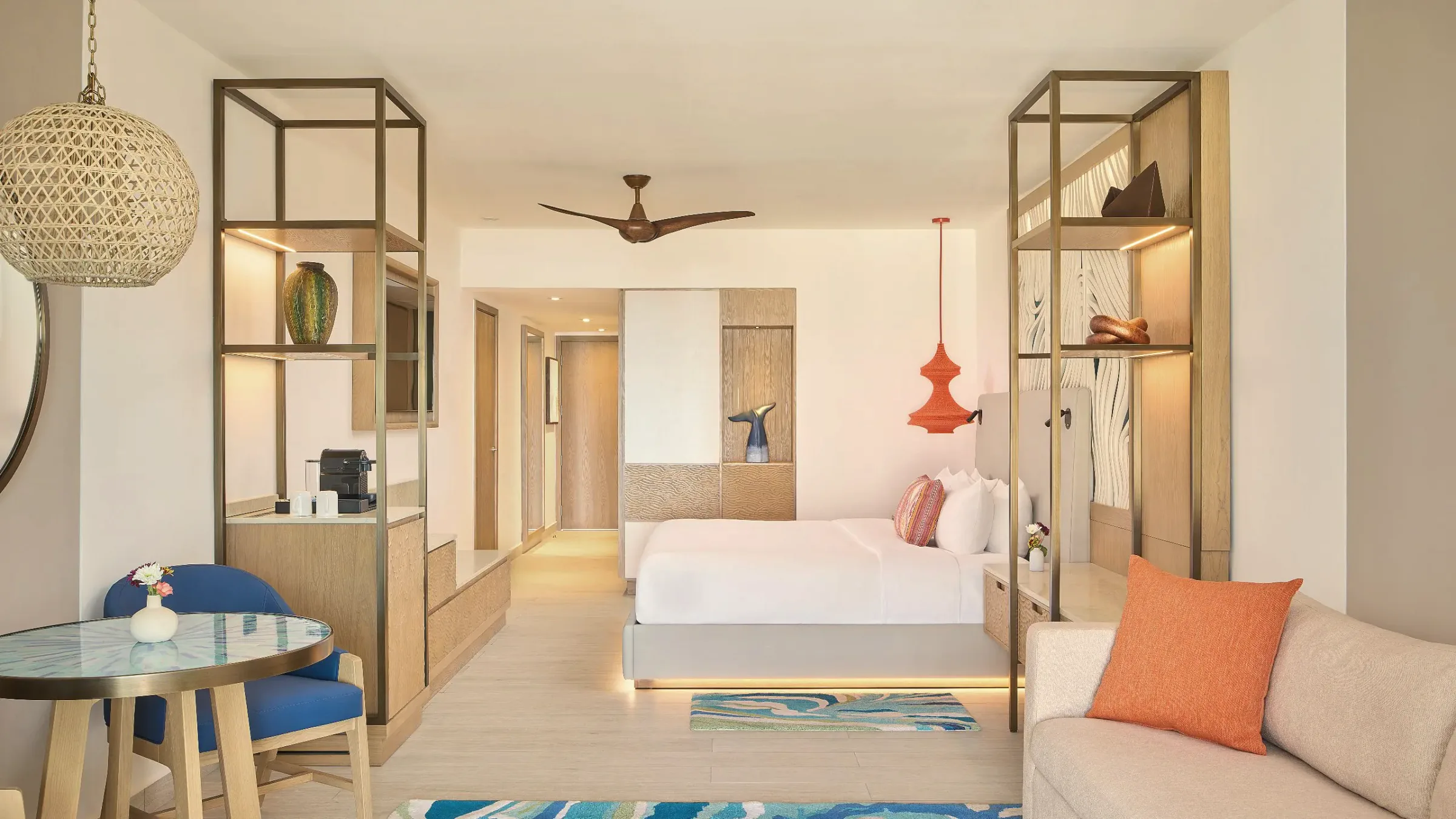 Guest room with a white bed, wood-framed shelving on both sides, a ceiling fan, a woven pendant lamp, a round table with a blue chair and a sofa with an orange cushion.