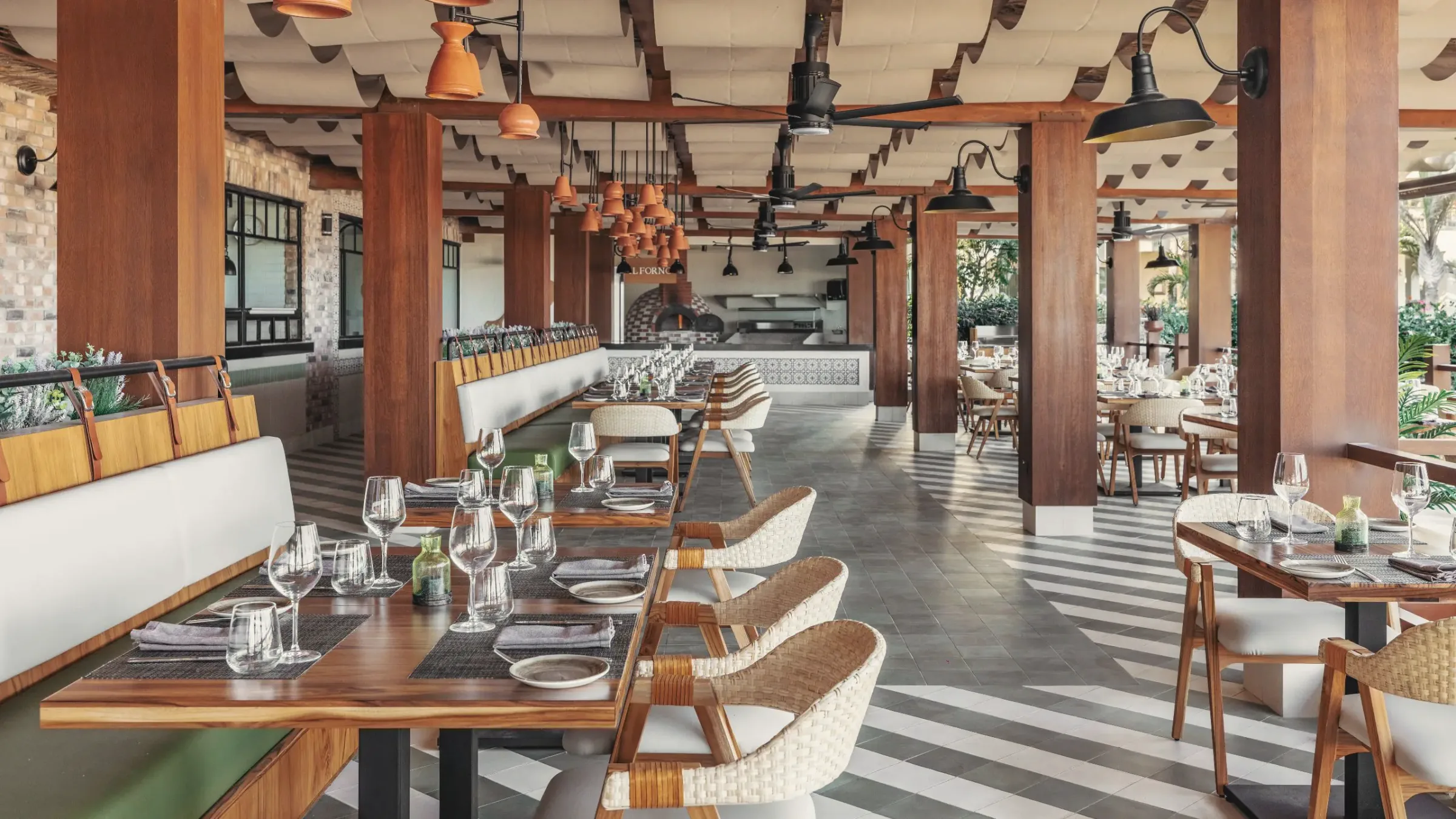 Restaurant dining room with wooden tables, woven chairs and white banquettes under a timber-beamed ceiling with terracotta pendant lamps, and an open kitchen with a wood-fired oven at the back.
