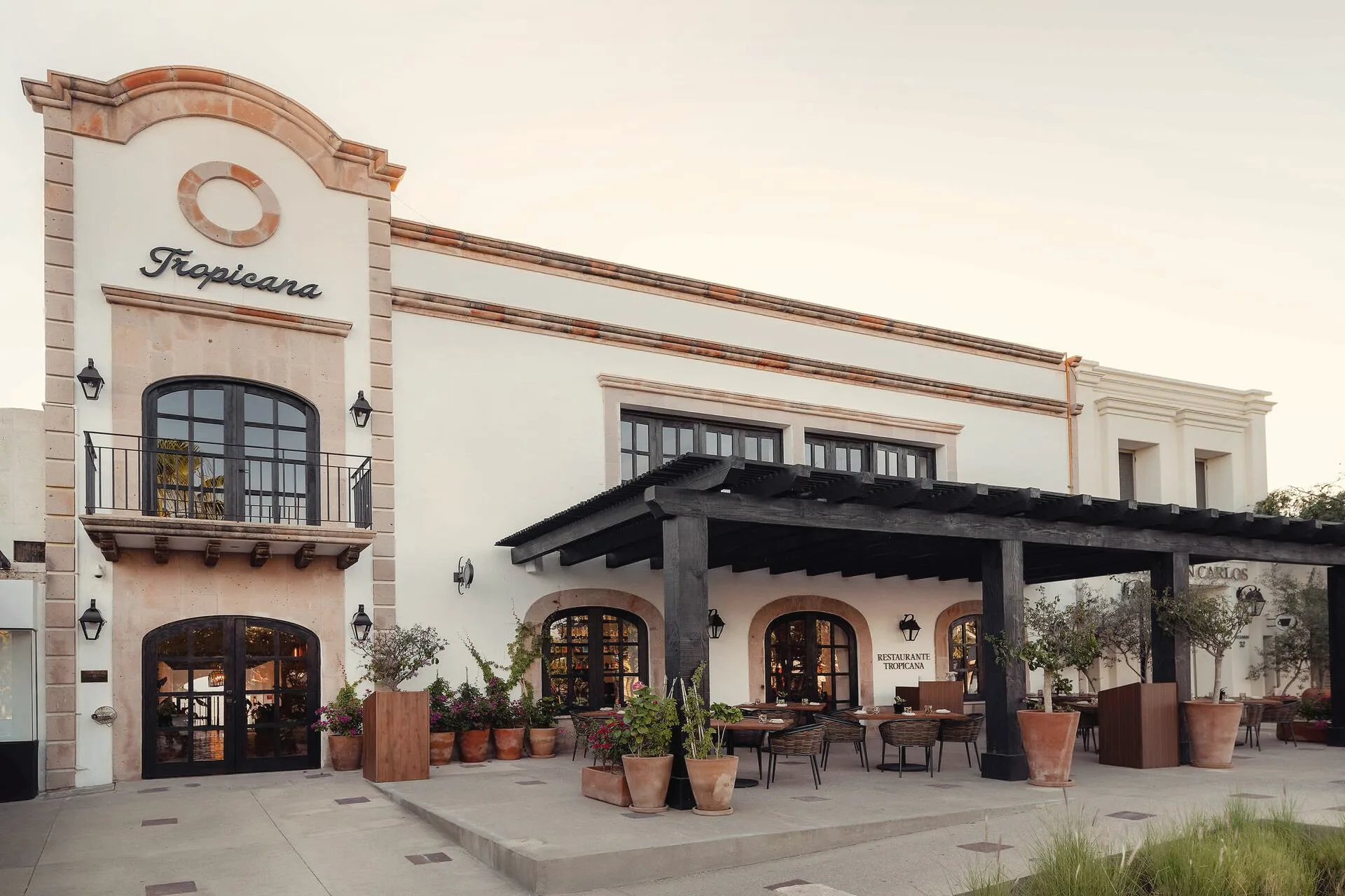 Front of the building in white and pink stone with a Tropicana sign, arched doors, a dark timber pergola and a restaurant terrace with potted plants.