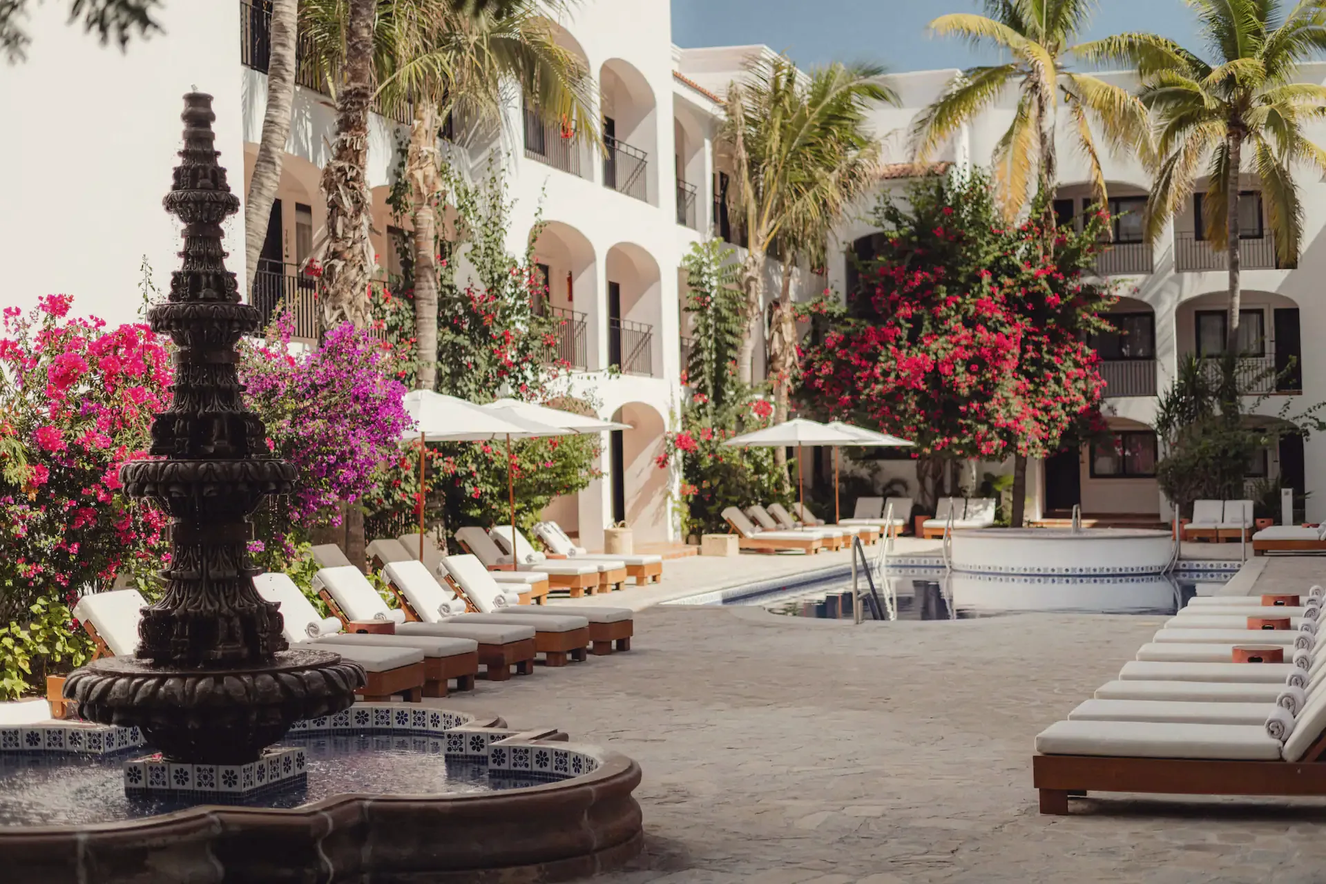 Courtyard with a tiered stone fountain, cushioned loungers and umbrellas by a pool, with bougainvillea and palms in front of white arched buildings.
