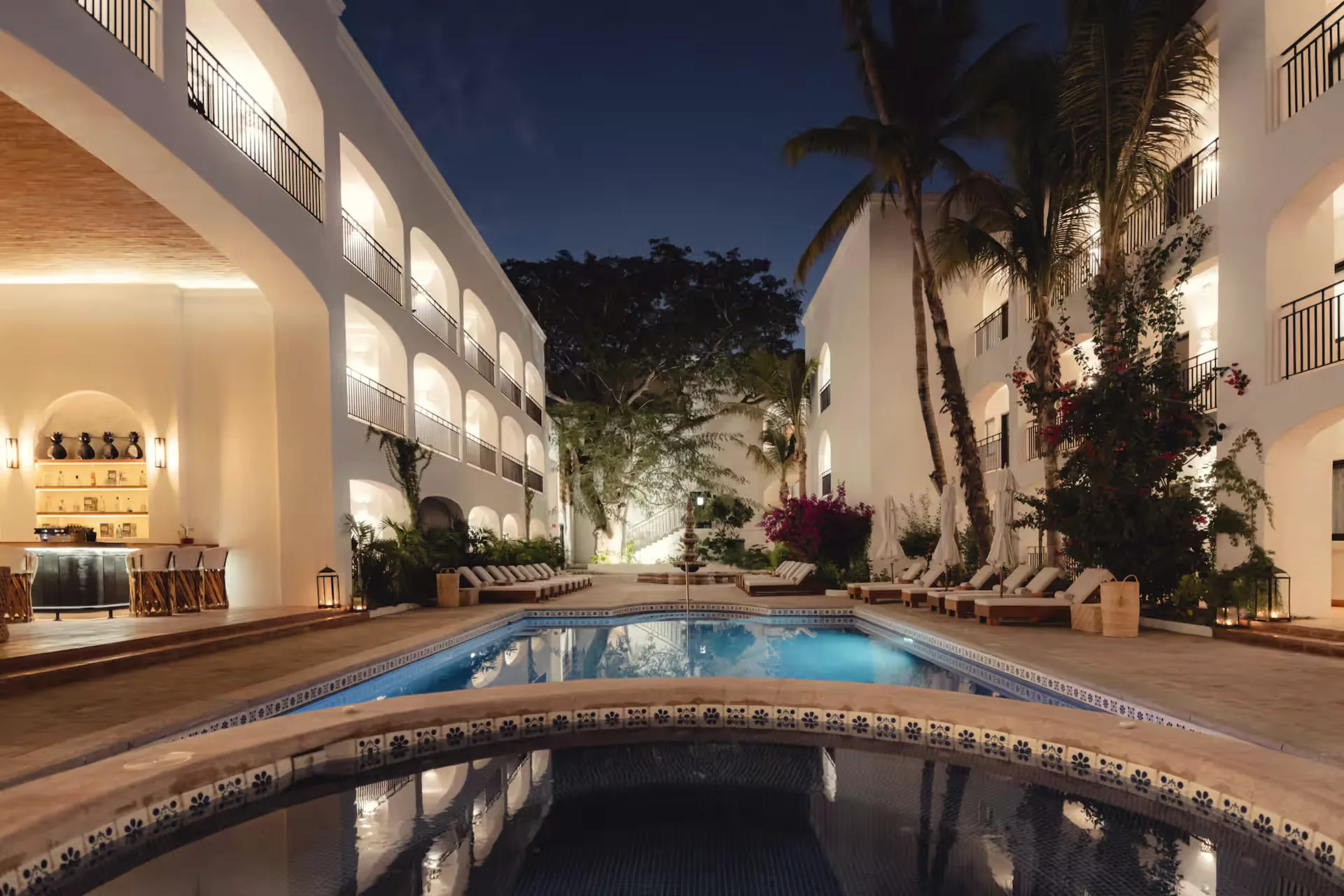 Courtyard pool at night with a tile-edged rim, lit arcaded building walls and palms against a dark blue sky.