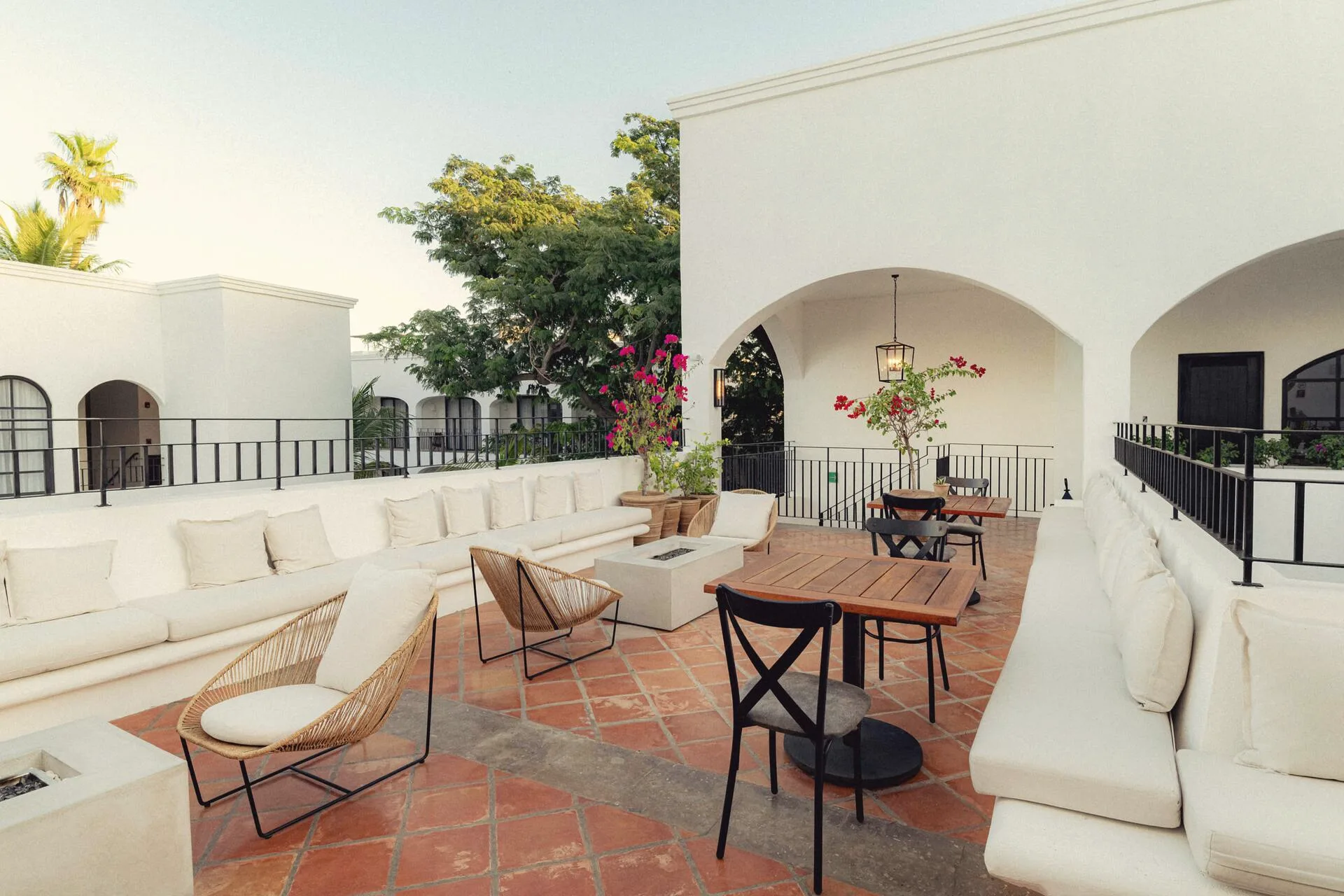 Rooftop terrace with white built-in sofas, rattan lounge chairs, a wooden dining table and terracotta tile floor, under white arches.
