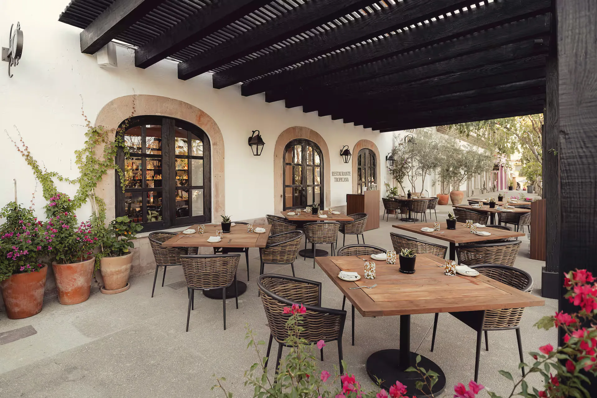 Covered restaurant terrace with wooden tables and woven chairs under a dark timber pergola, with flowering plants and arched windows.