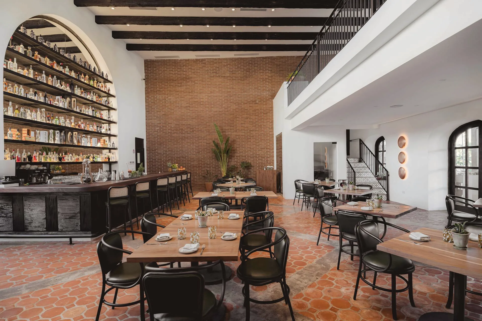 Restaurant interior with a tall wall of bottles behind a bar, wooden tables, black chairs and a terracotta floor.