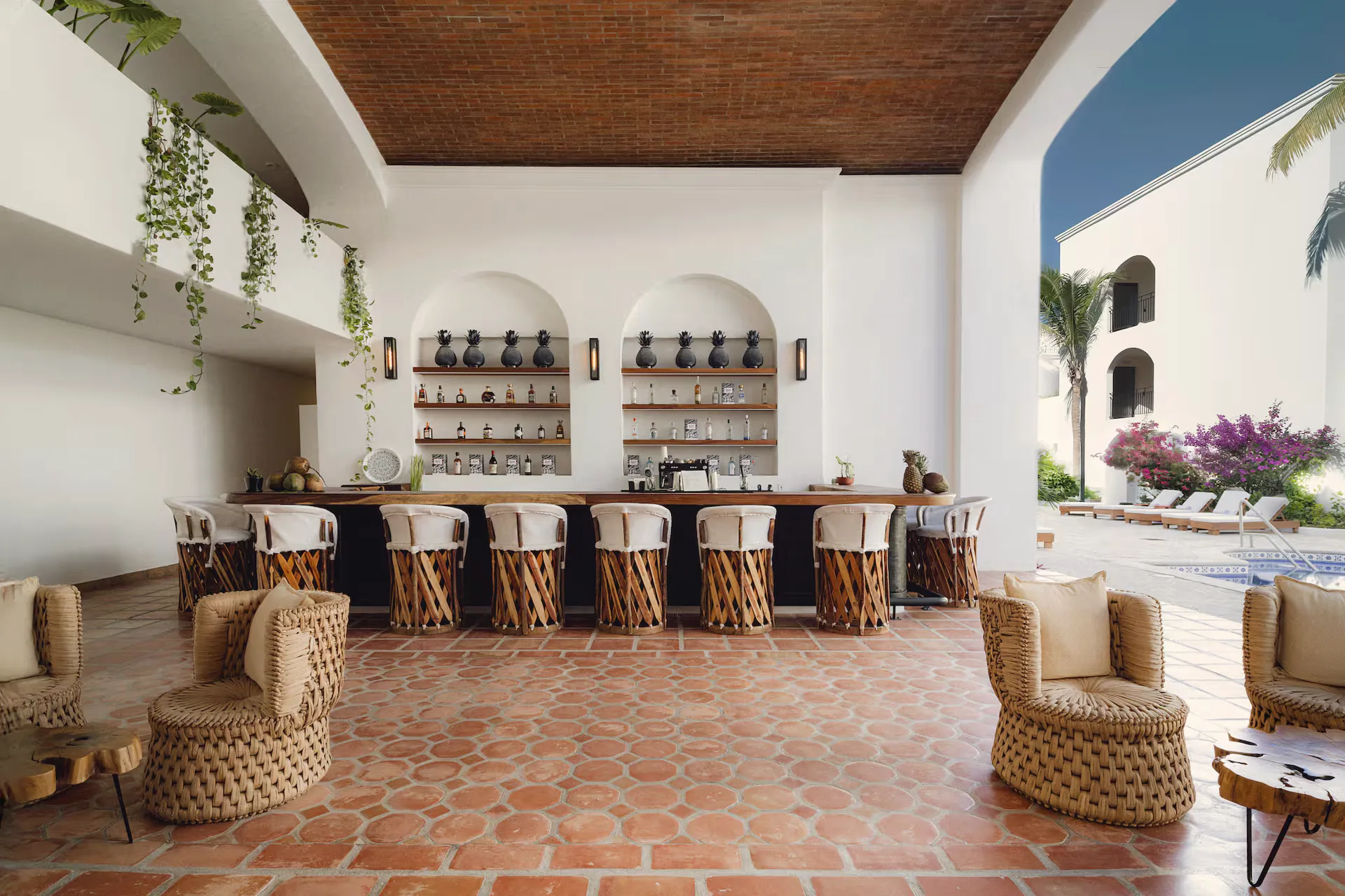 Open-air bar with a row of leather-strapped stools, arched shelves holding pottery and a terracotta floor, with the pool visible beyond.