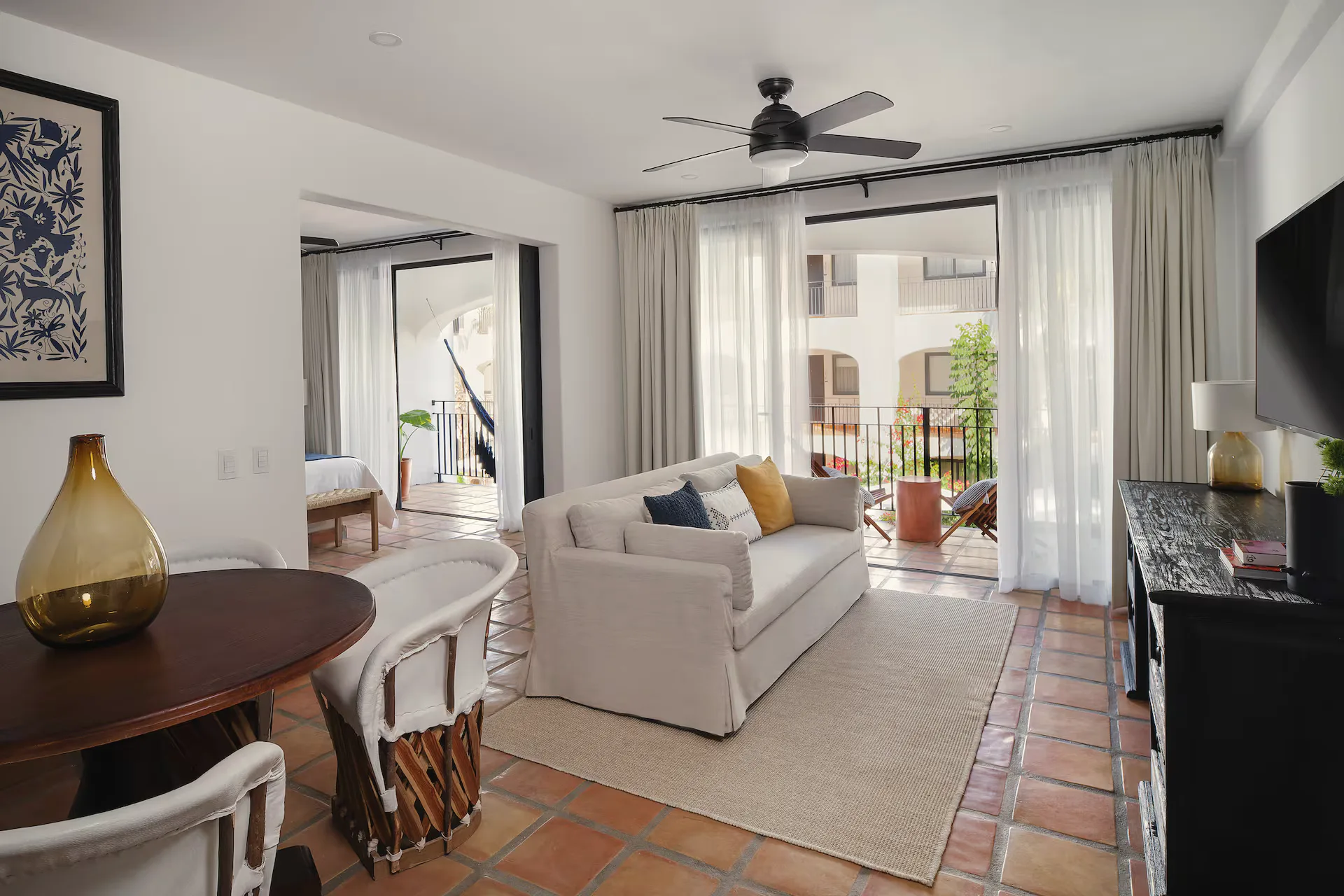 Living room with a white sofa, a rug, a round wooden table with leather-and-wood chairs, a ceiling fan and sheer curtains onto a balcony.