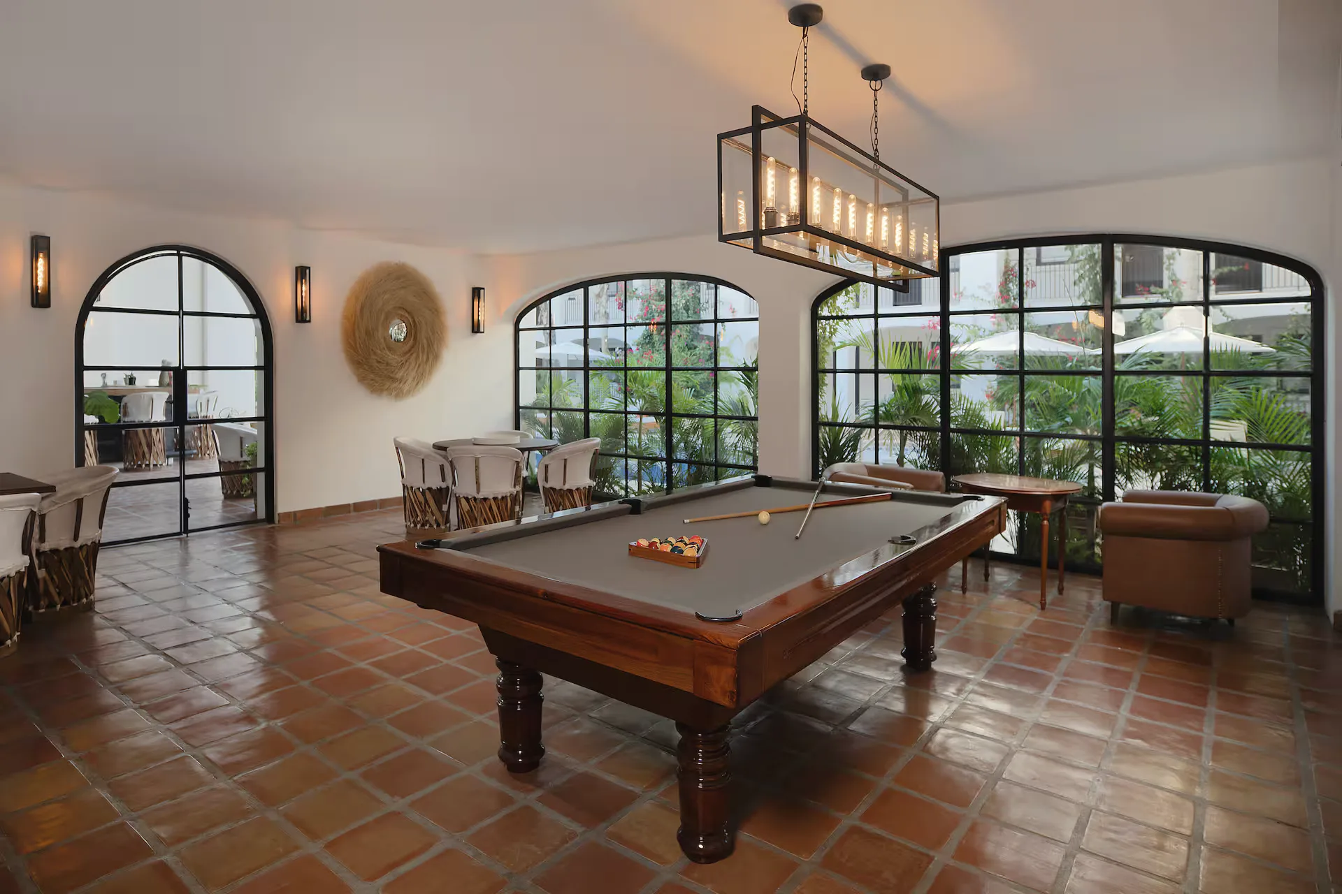 Games room with a billiard table, a long pendant lamp, tall black-framed arched windows and a terracotta tile floor.