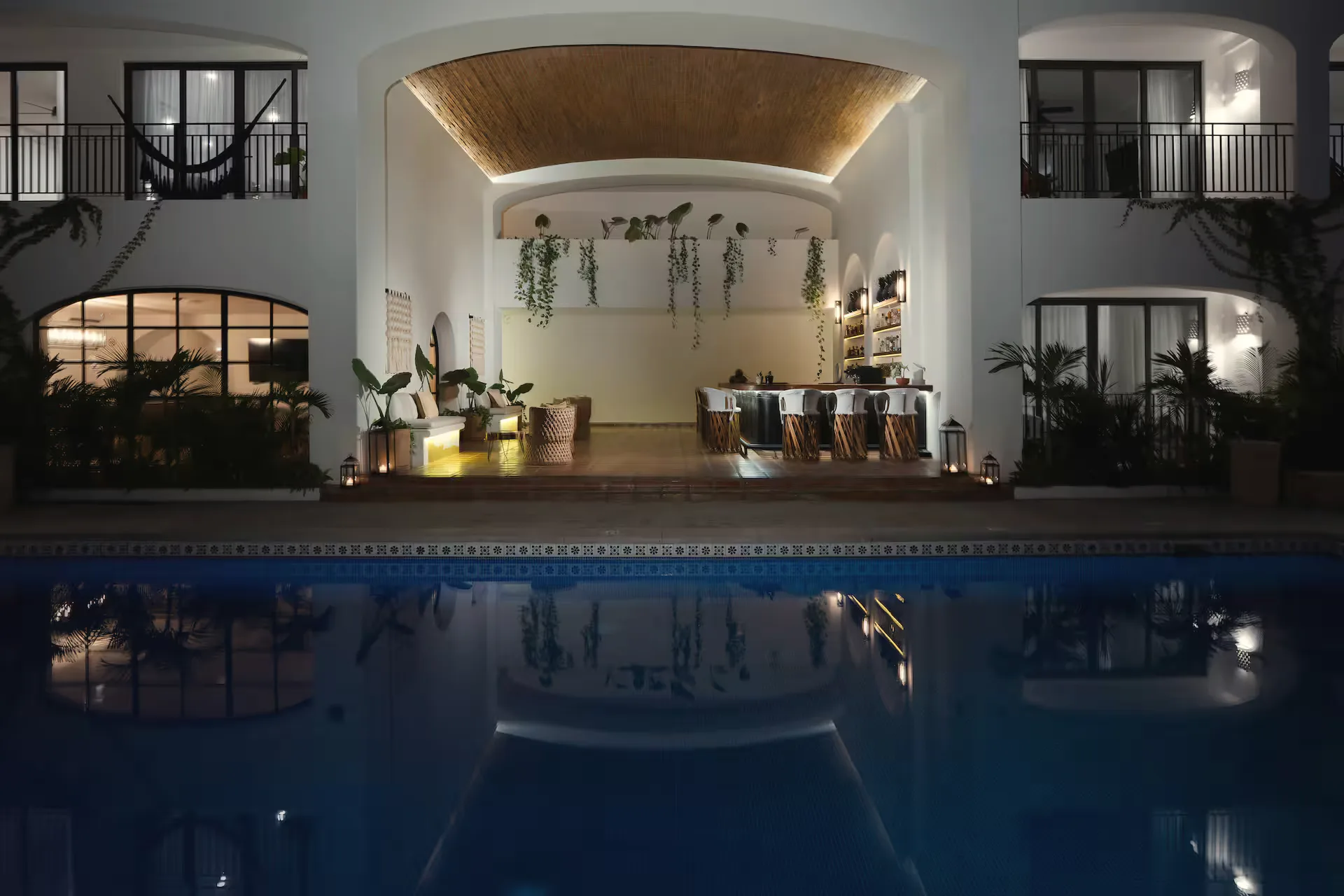 Dark pool at night in front of a lit arched terrace with hanging plants and a dining table.