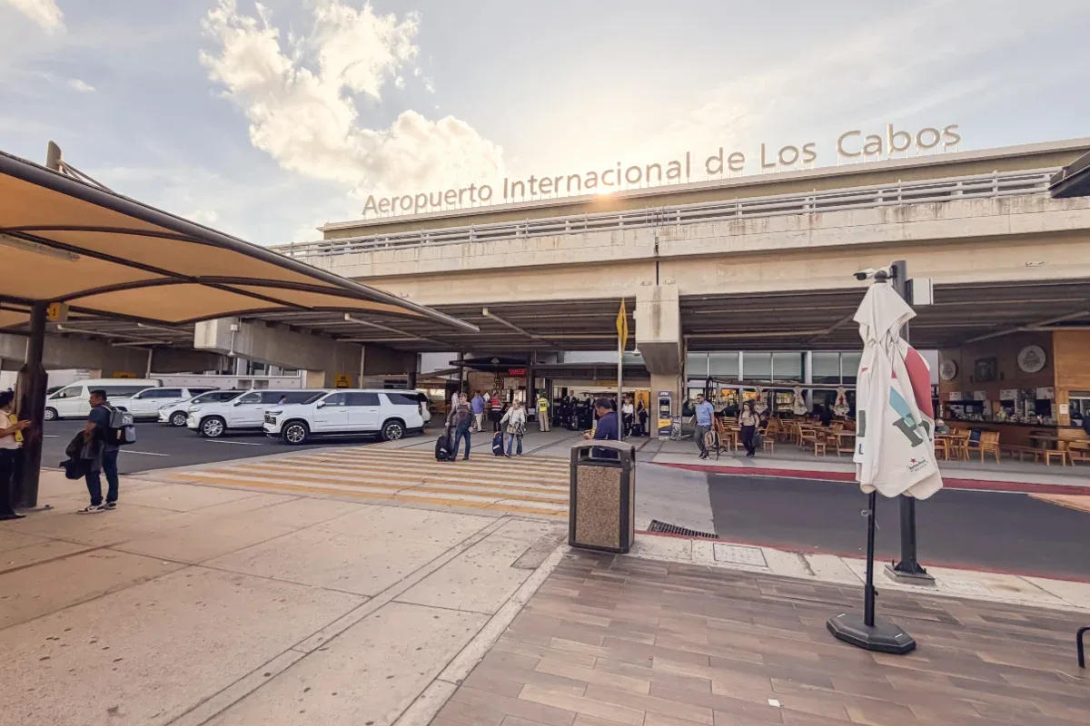 The arrivals curb at Los Cabos International Airport under the terminal sign, with white vans waiting and travelers with luggage