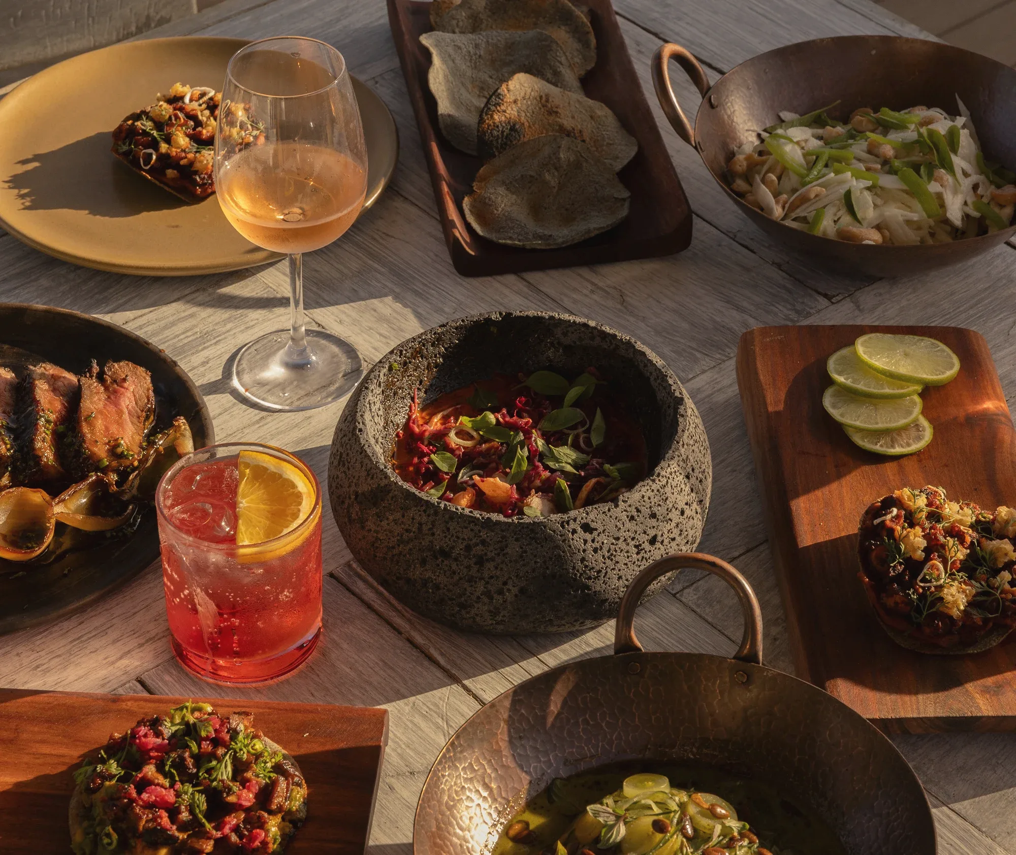 A sunlit table of shared dishes: a rosé, a red cocktail with an orange slice, tortillas, a stone bowl of ceviche and sliced steak